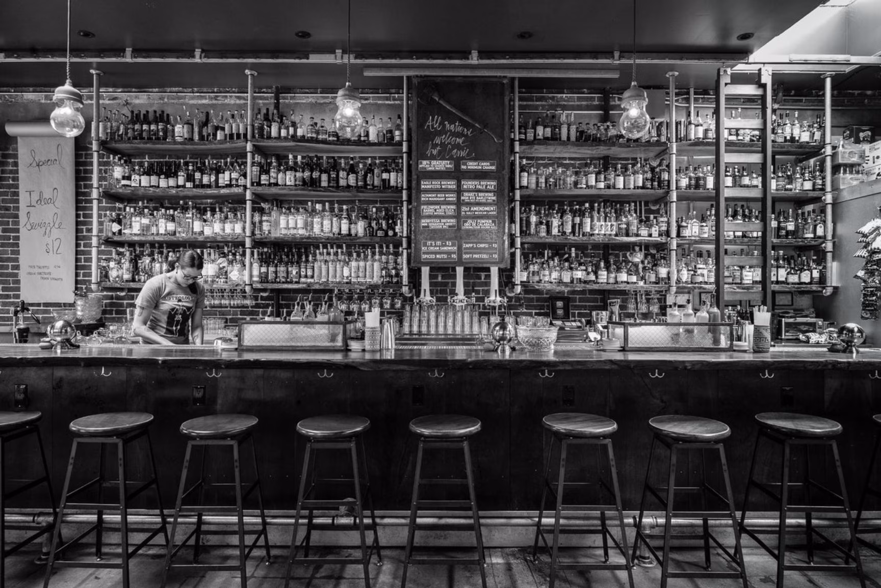 Black and white photo of a bartender behind a fully stocked bar.