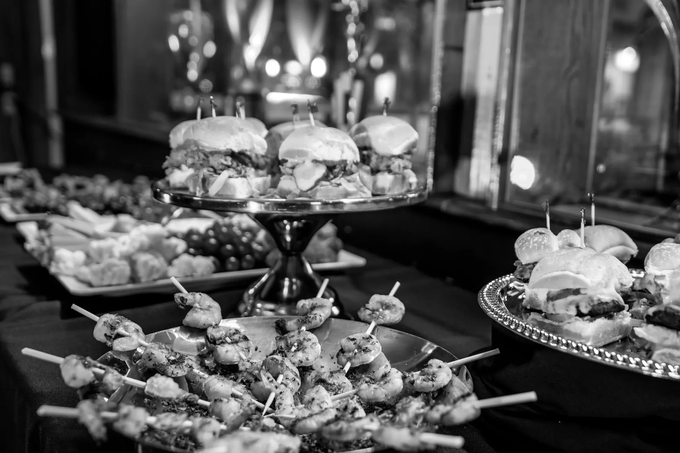 Monochrome photo of sliders, shrimp skewers, and various appetizers on trays.
