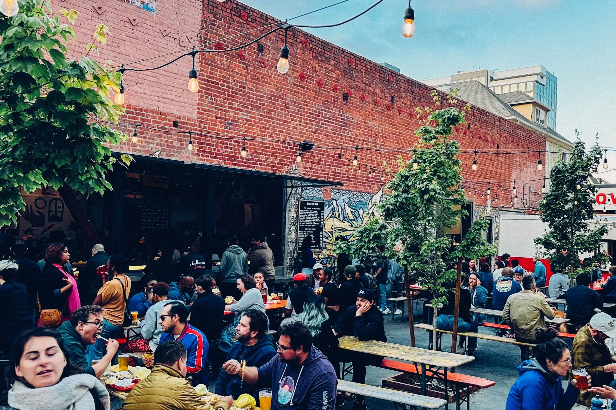 Outdoor beer garden with many people, string lights, and trees.