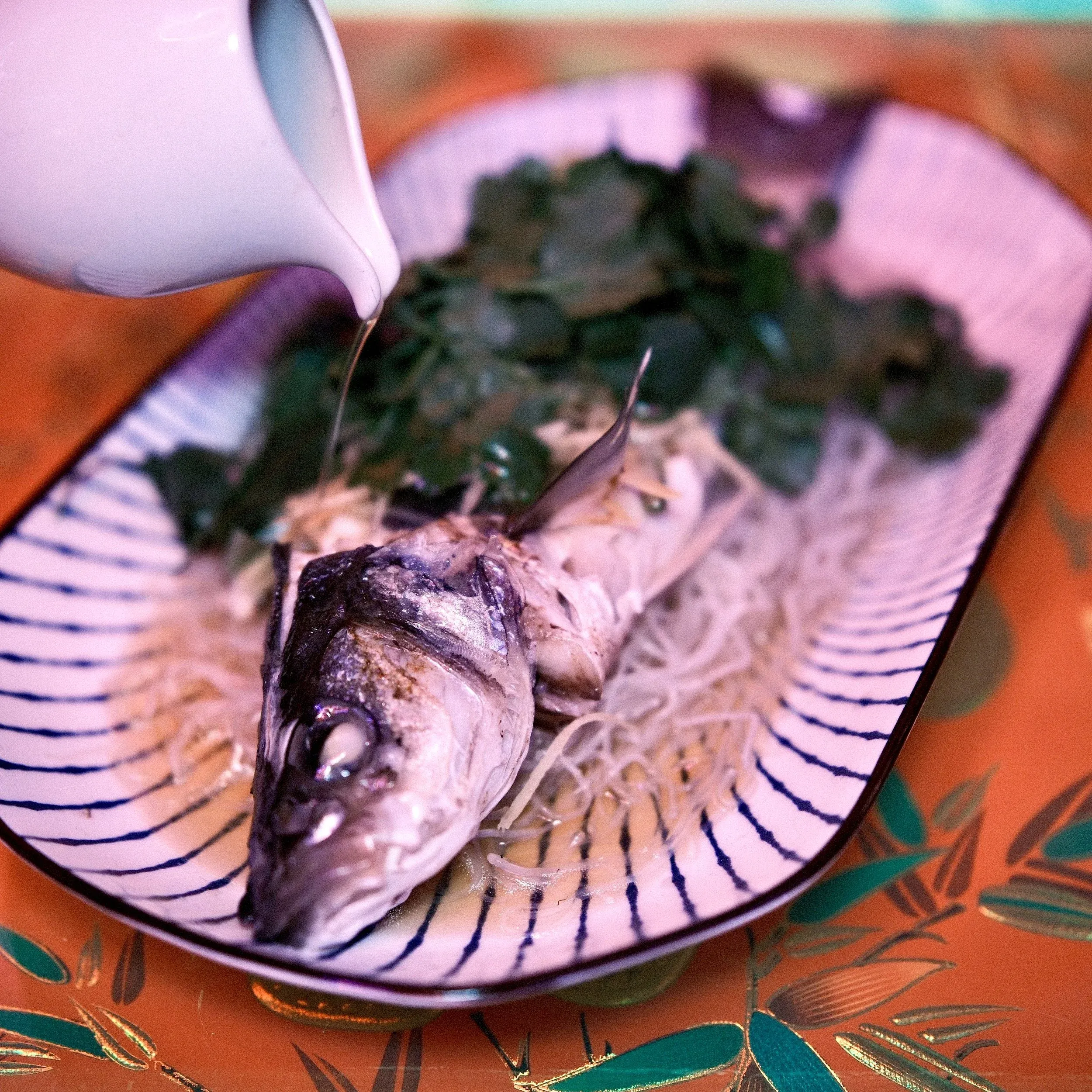 White pitcher pours liquid over whole cooked fish and herbs.