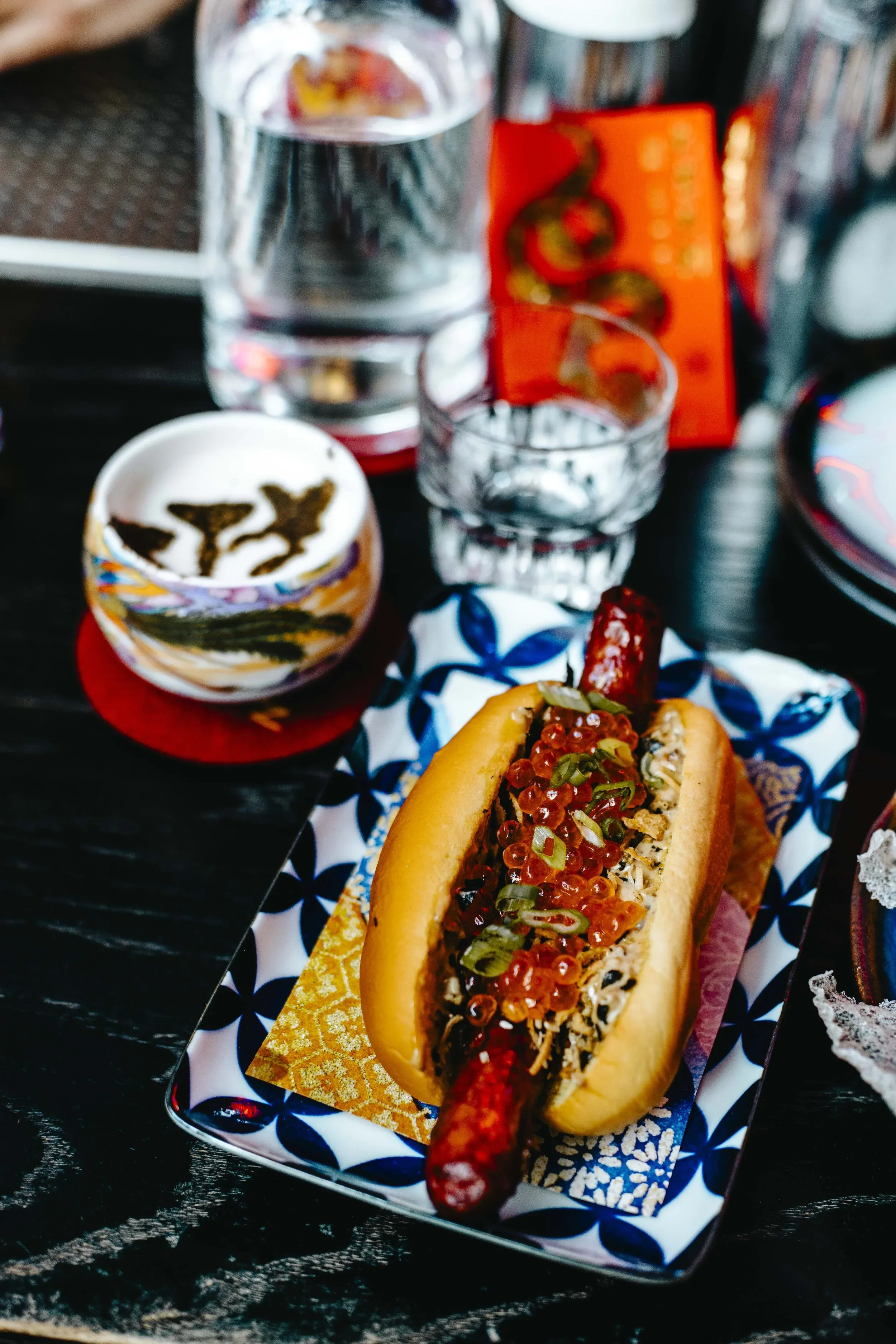 Hot dog topped with red roe and green onions on a patterned plate.