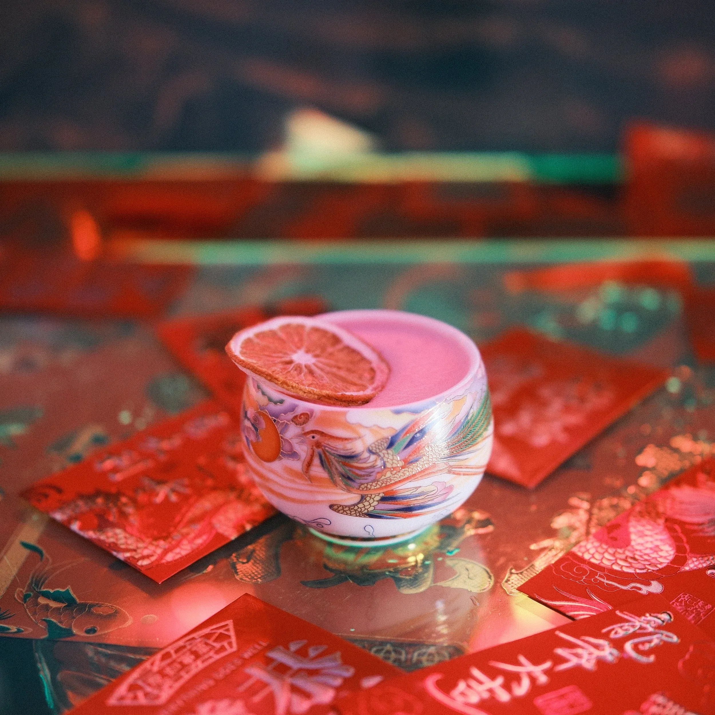 Pink drink in phoenix cup with dried citrus slice and red envelopes.