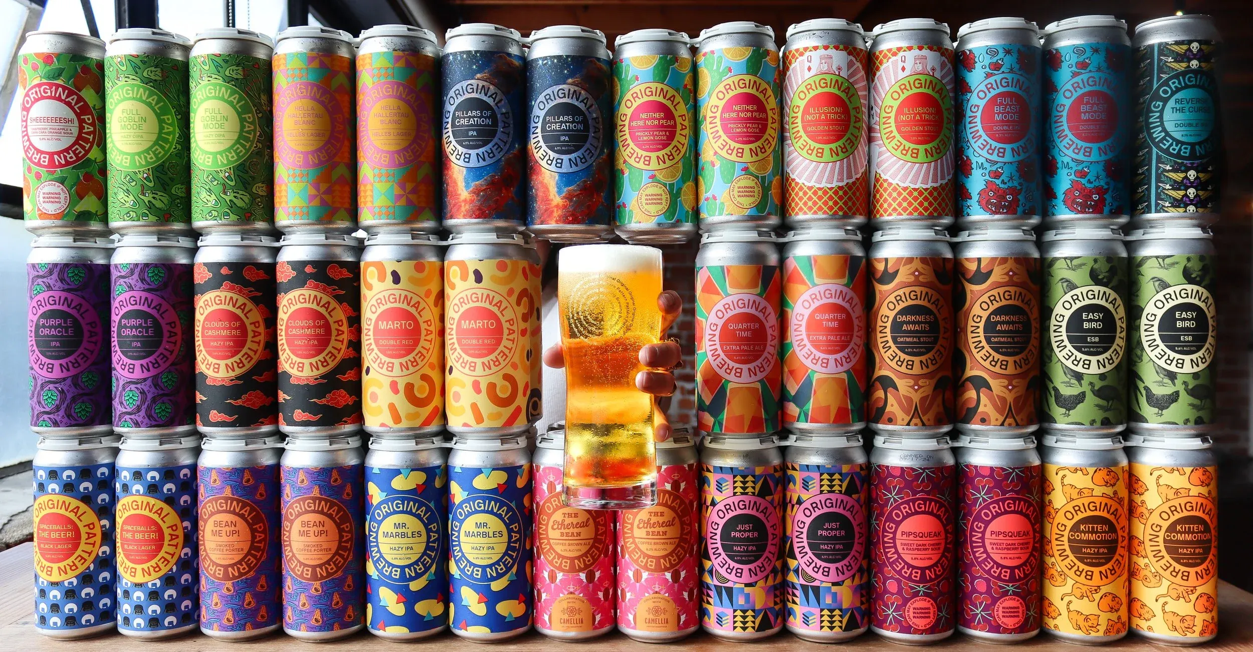 Colorful Original Pattern Brewing beer cans stacked, with a hand holding a glass.
