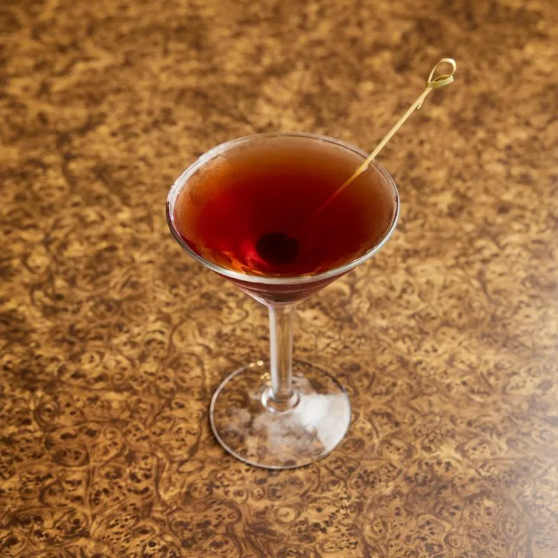 Dark red cocktail in a martini glass with cherry on a patterned surface.