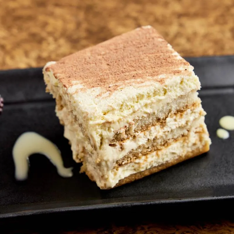 Slice of tiramisu with cocoa powder on a black plate.