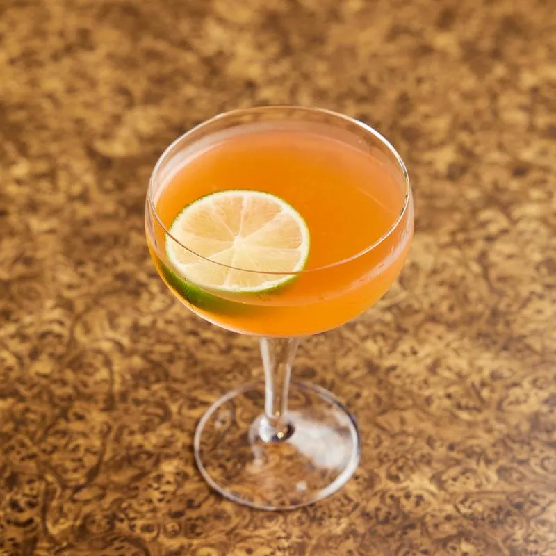 Orange cocktail in coupe glass with lime slice on patterned table.