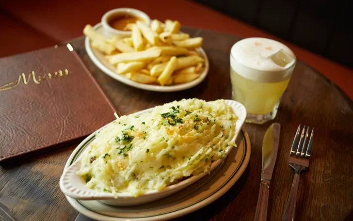 Mashed potato-topped dish, fries, cocktail, menu, on wooden table.