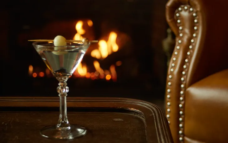A martini with an olive in front of a fireplace and leather chair.