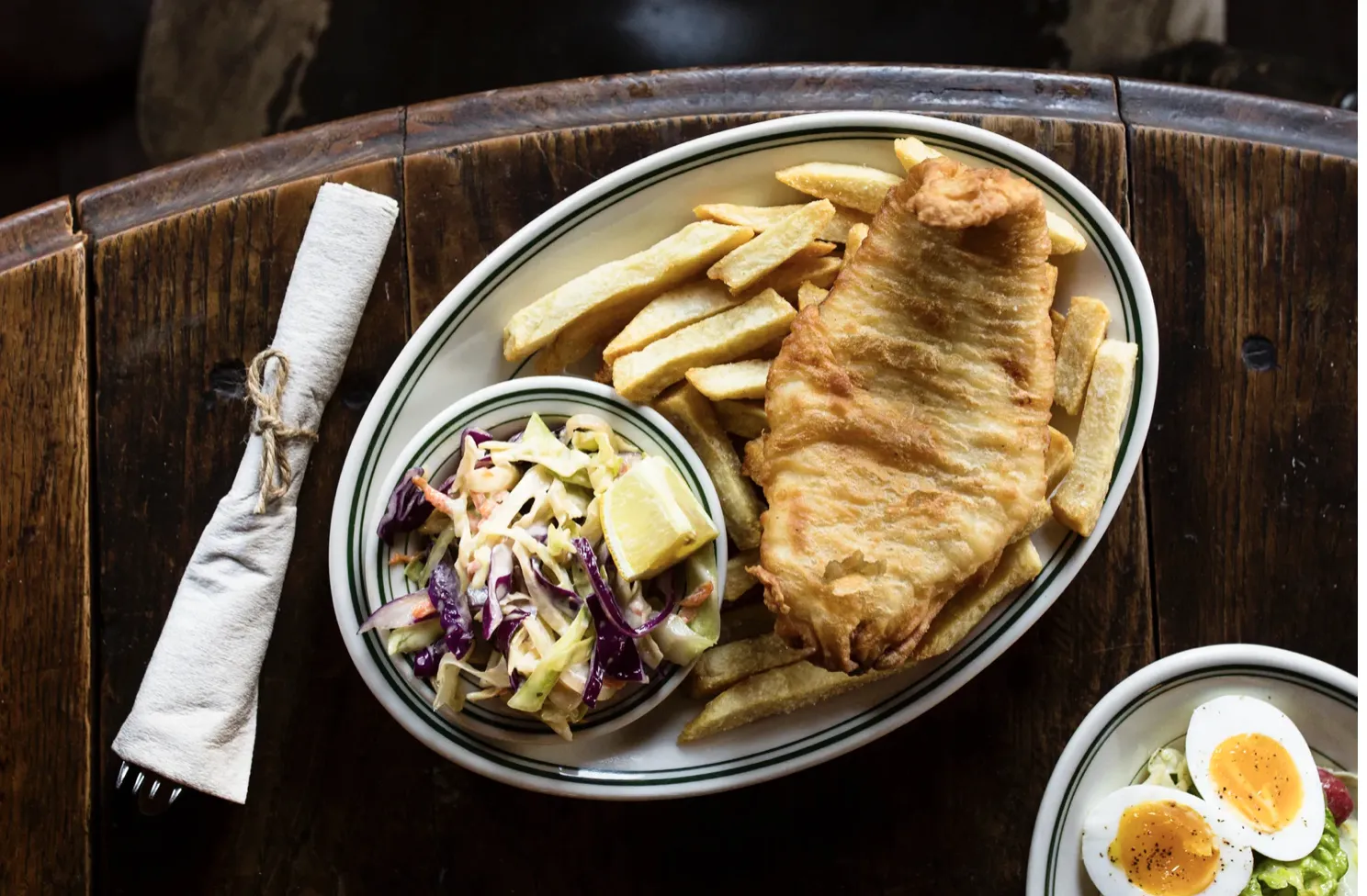 Plate of fish and chips, coleslaw, and napkin on a wooden table.