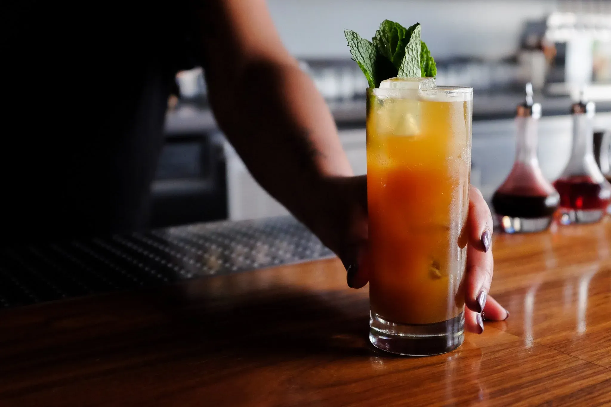 Hand holding an orange cocktail with mint garnish on a bar.