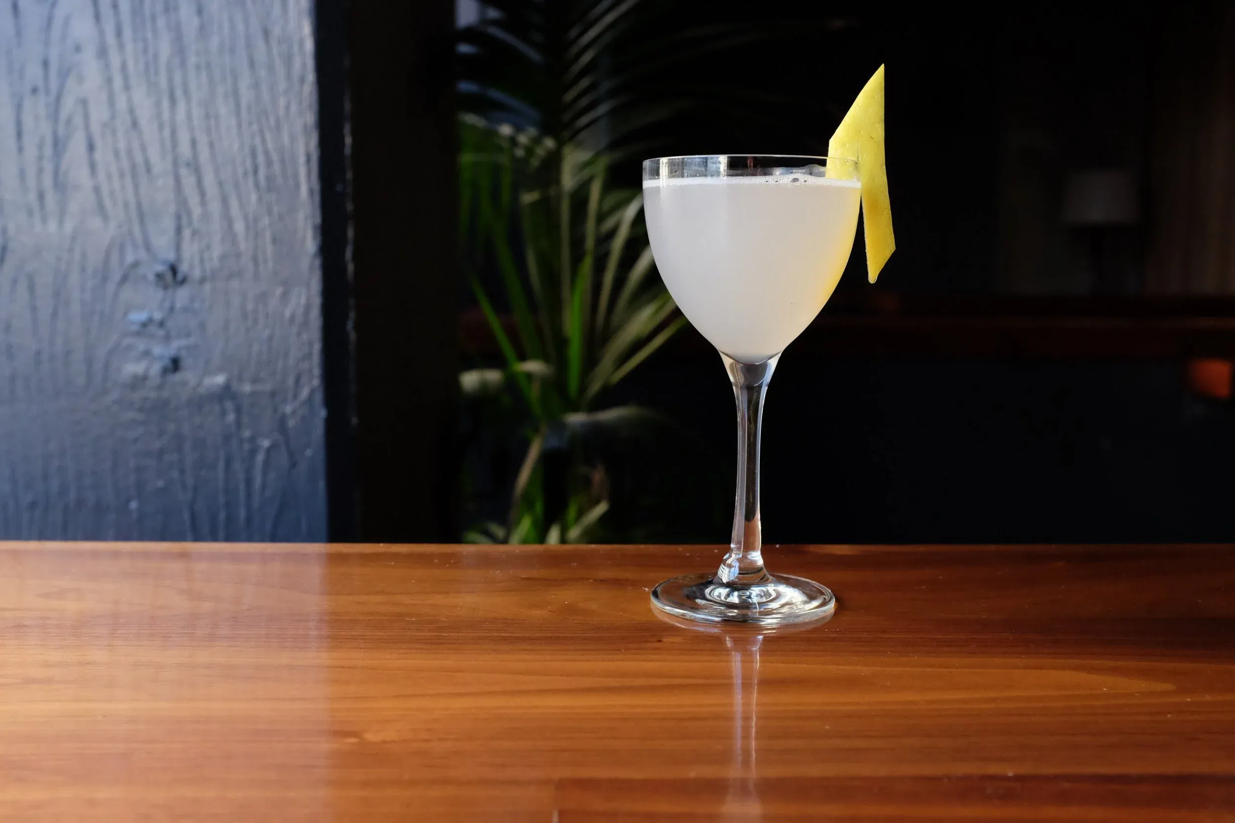 Pale cocktail with citrus peel garnish on a polished wooden table.