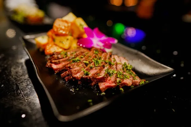 Sliced steak, roasted potatoes, and orchid flower on a dark plate.
