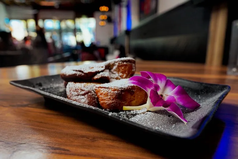 Black plate with fried pastries, powdered sugar, and a purple orchid.
