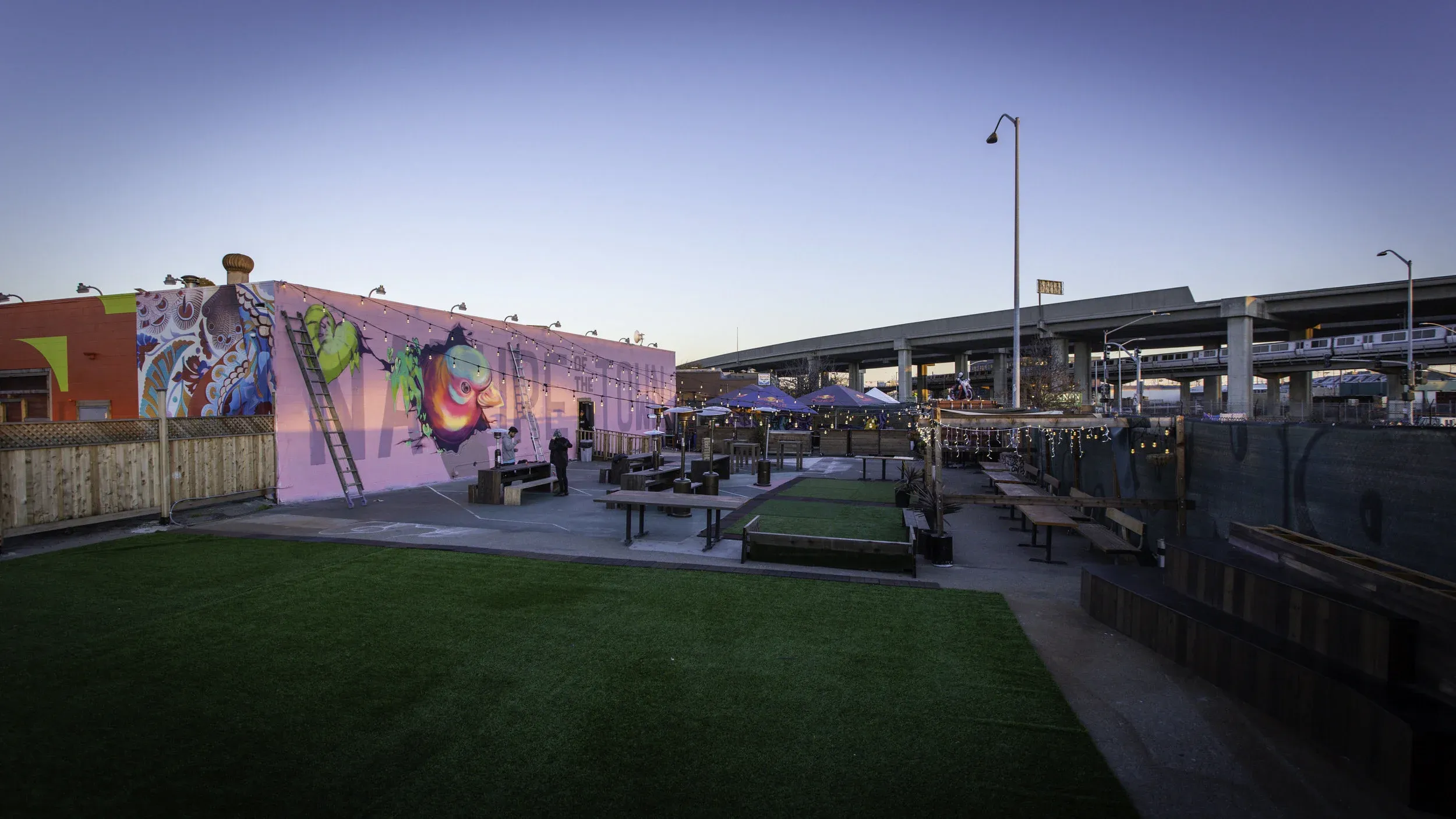 Outdoor patio with colorful mural, string lights, and elevated highway.