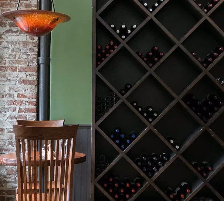 Wine bottles in a large rack beside a table and brick wall.