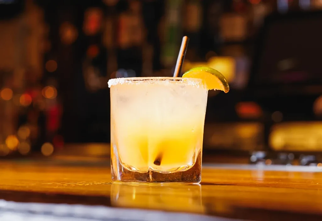 Margarita with a salted rim, lime, and straw on a wooden bar.