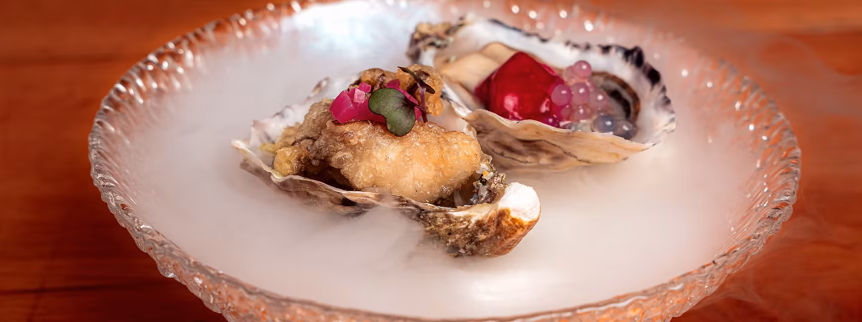 Two oysters, one fried, one with red elements, served on a smoky dish.
