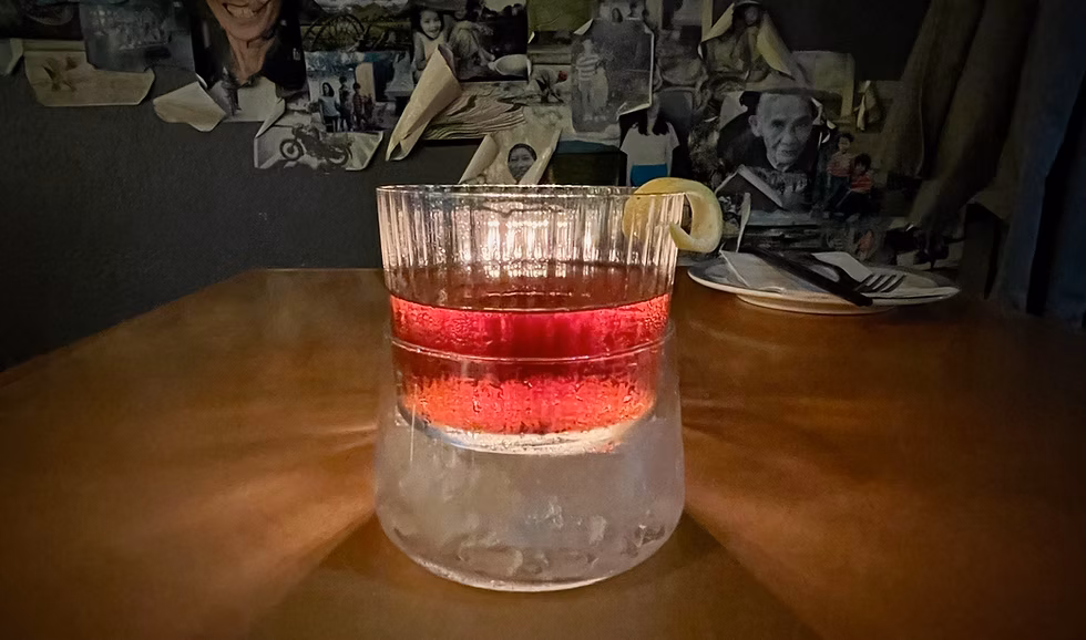 Red drink in ribbed glass with ice, lemon peel garnish.