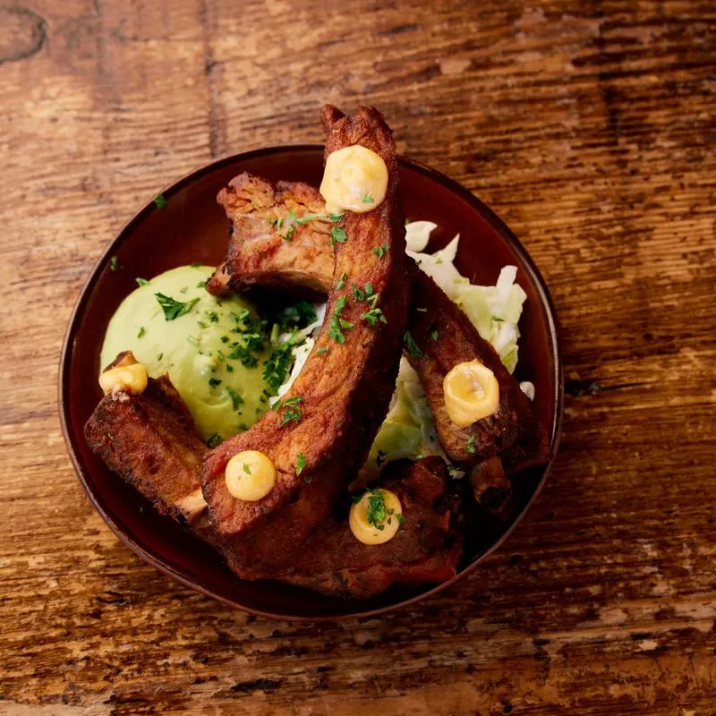 Brown plate of ribs with green and yellow sauces on a wood table.