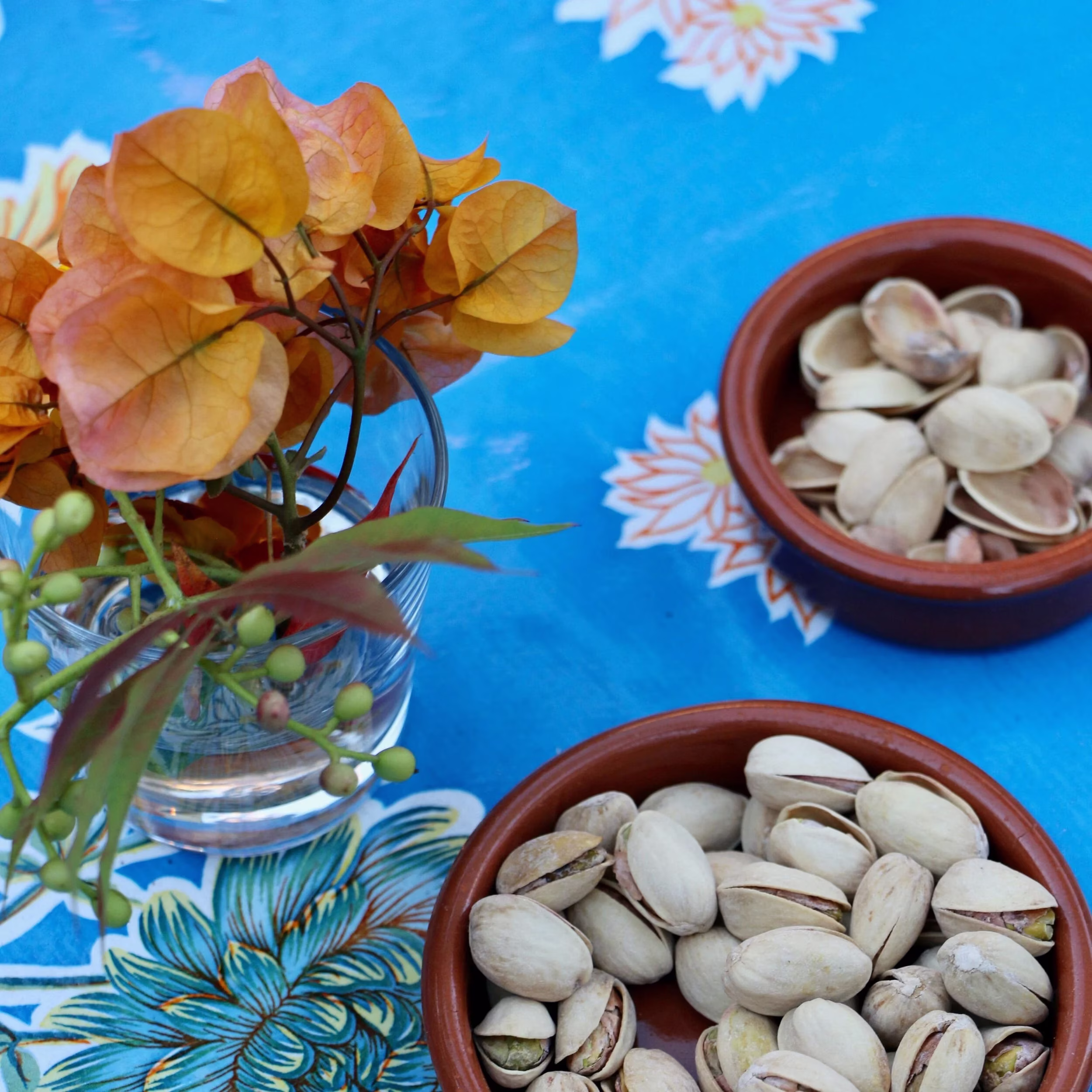 Orange flowers, green berries, and two bowls of pistachios on a blue tablecloth.