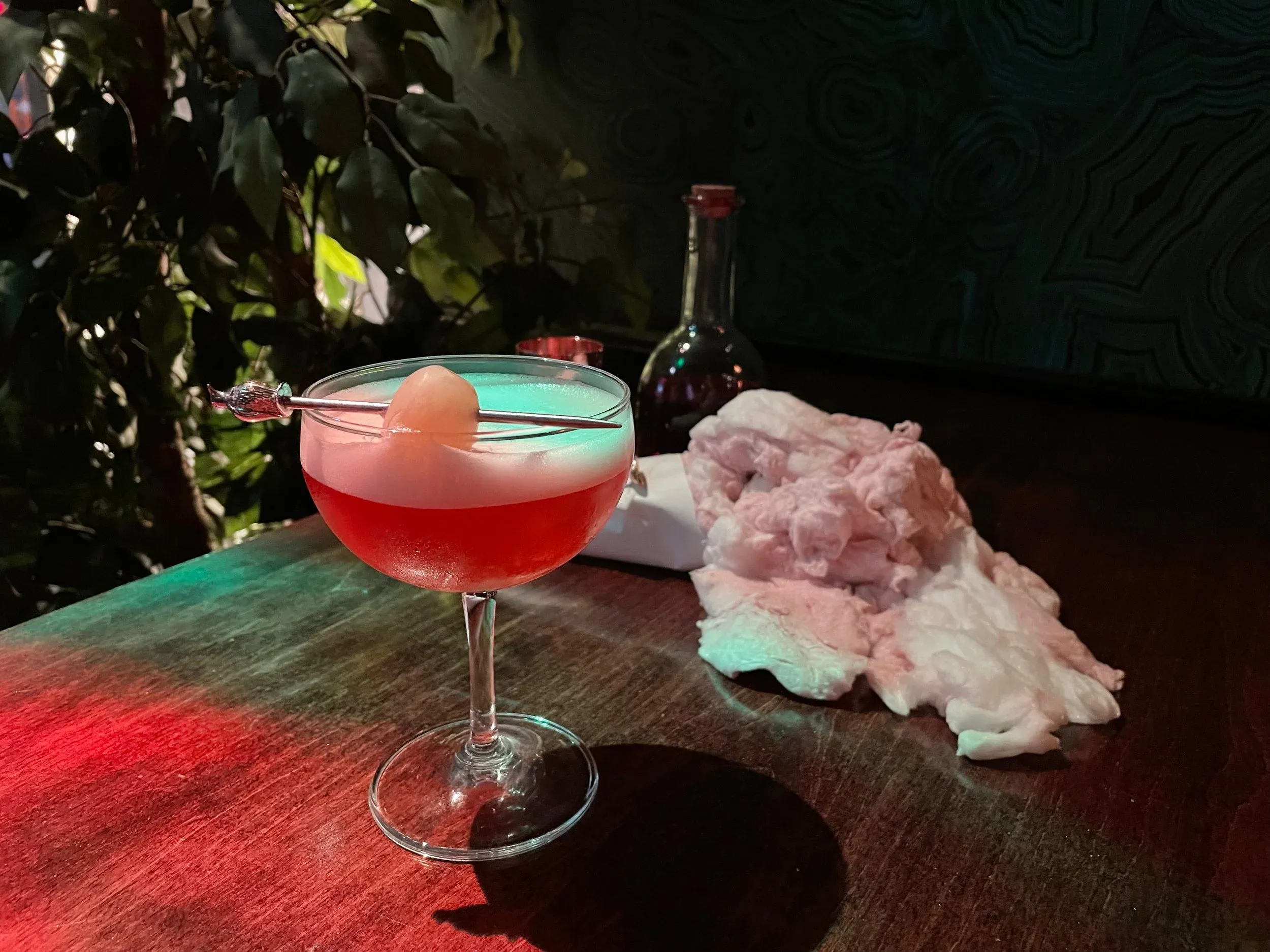 Red cocktail in coupe glass with foamy top and fruit garnish.