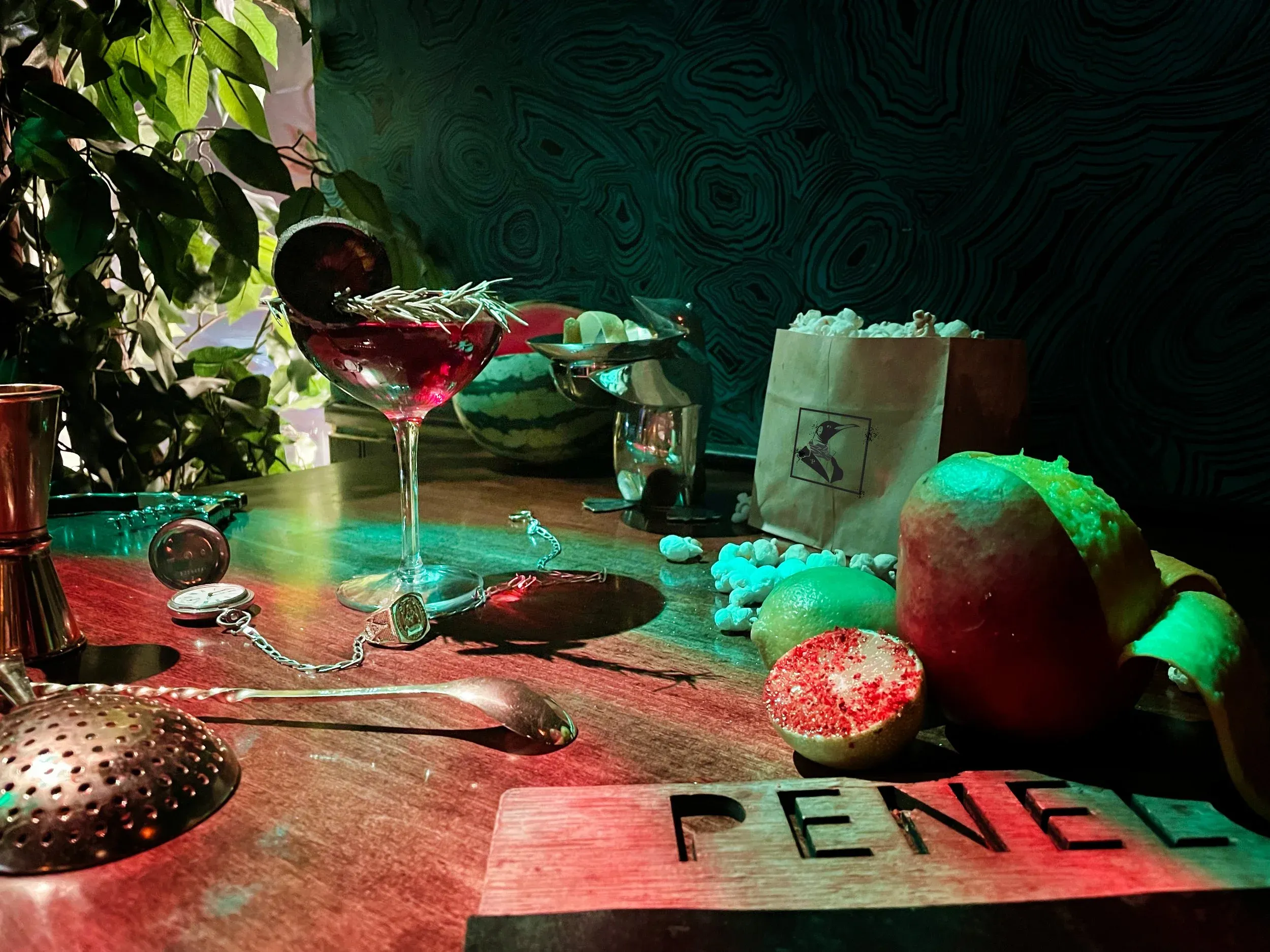 Red cocktail, bar tools, fruit, popcorn, and a watch on a table.