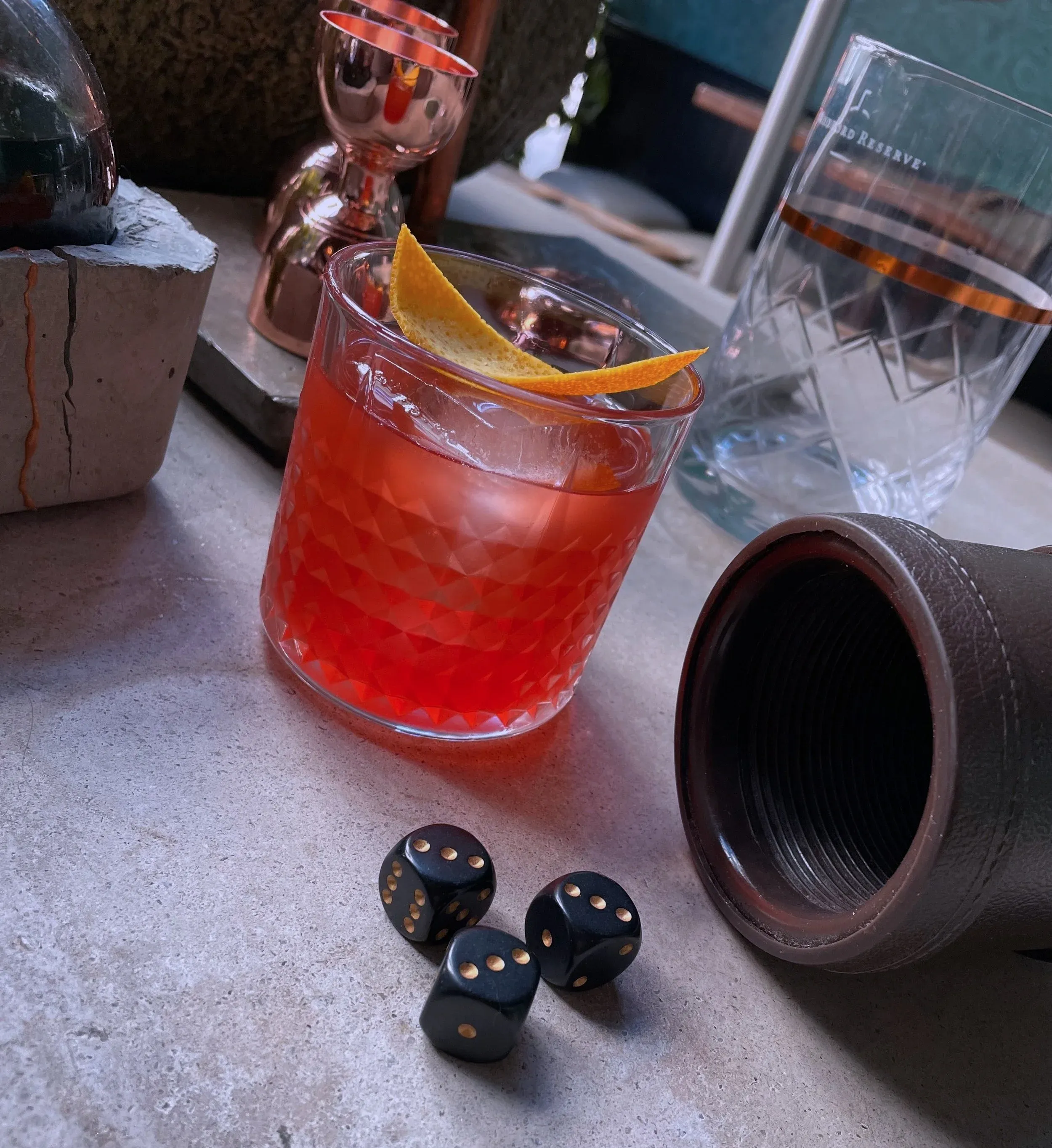 Red cocktail with orange peel, dice, and barware on a gray surface.