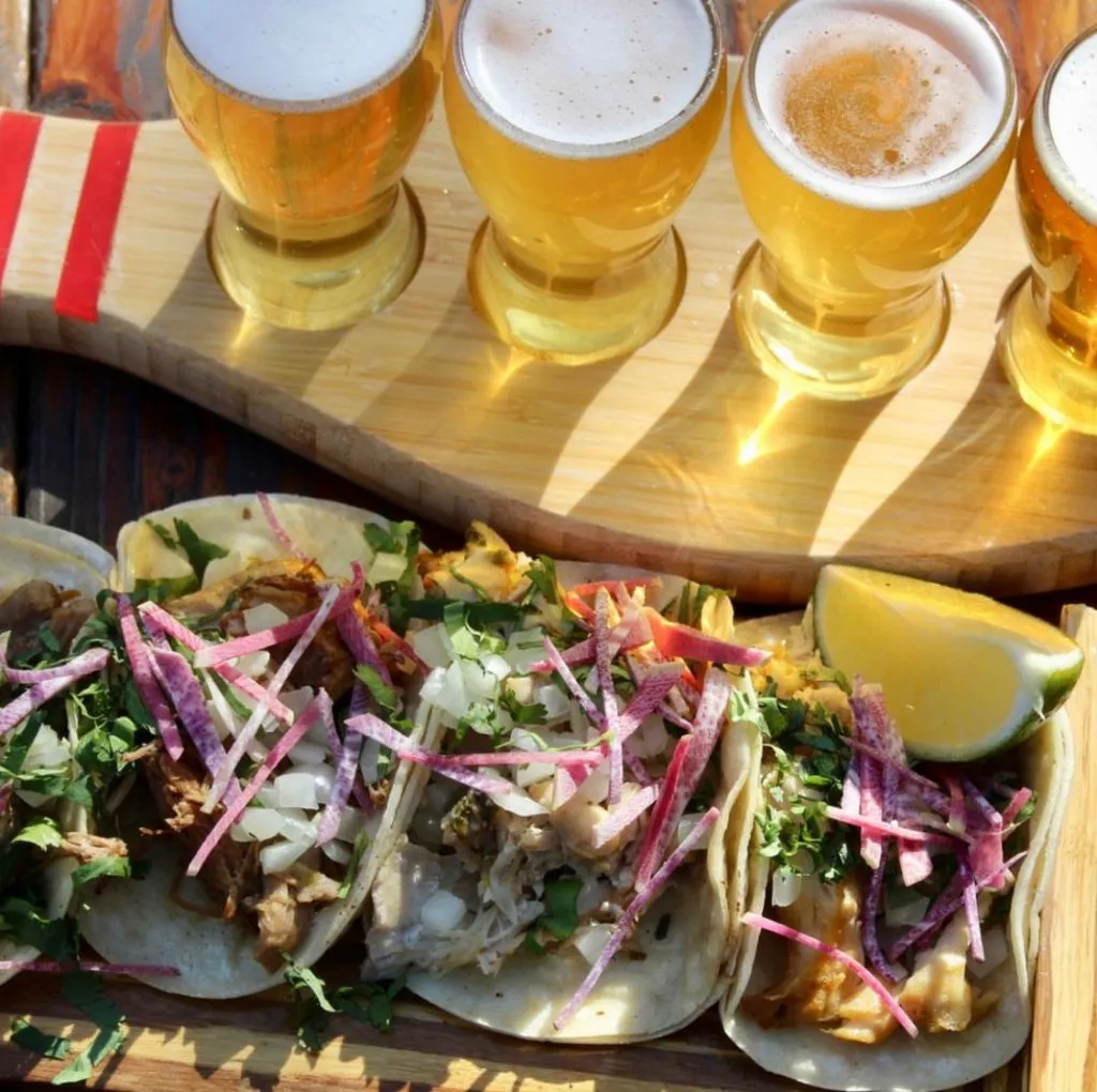 Tacos with colorful toppings and a beer flight on a wooden board.