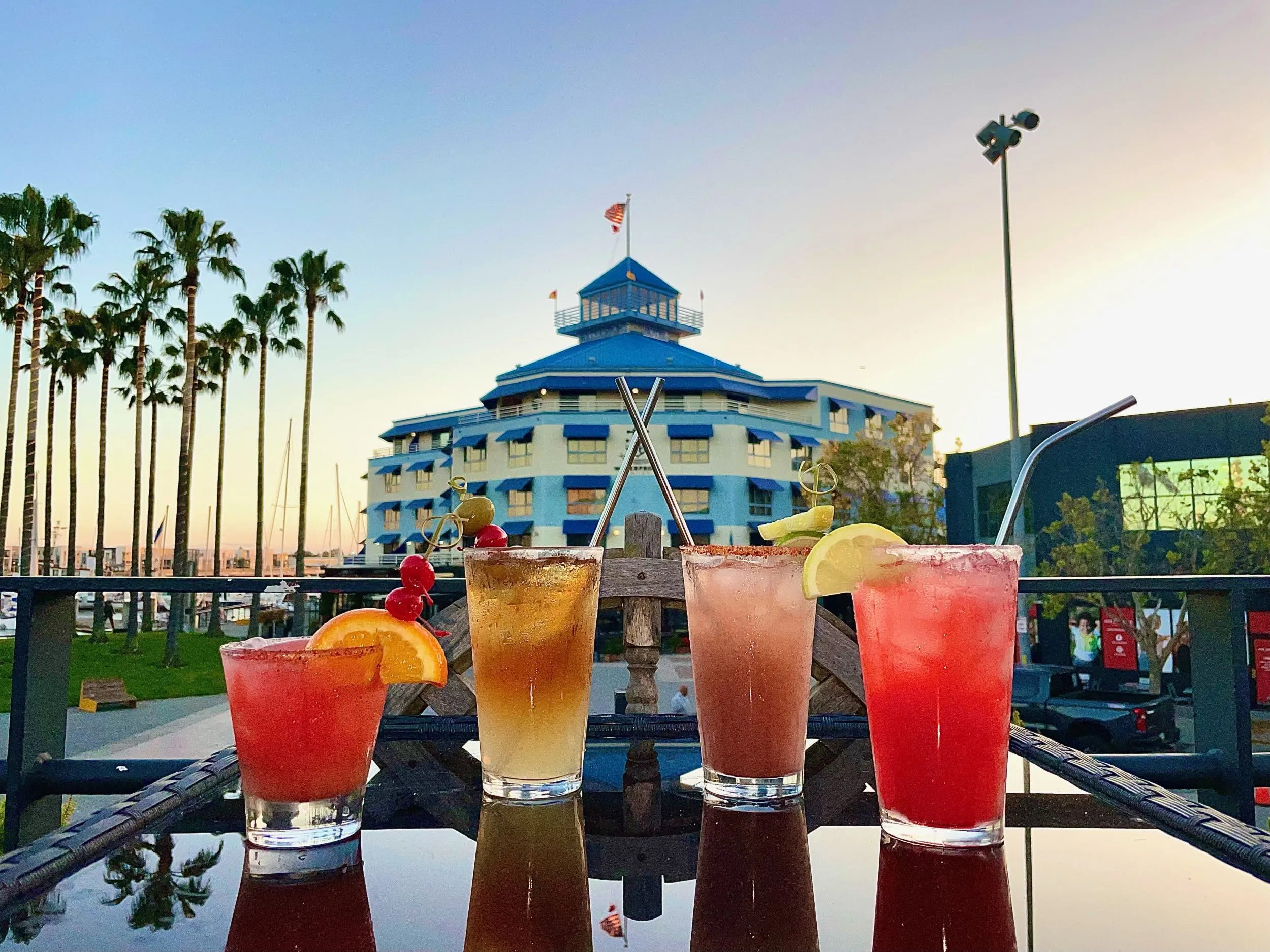 Four colorful cocktails on a table overlooking a marina at dusk.