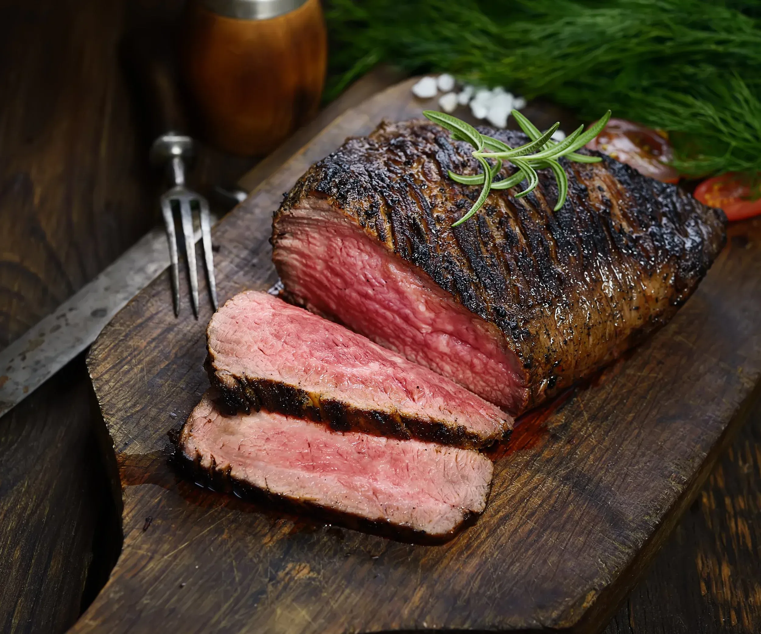 Sliced medium-rare beef, garnished with rosemary on a wooden board.