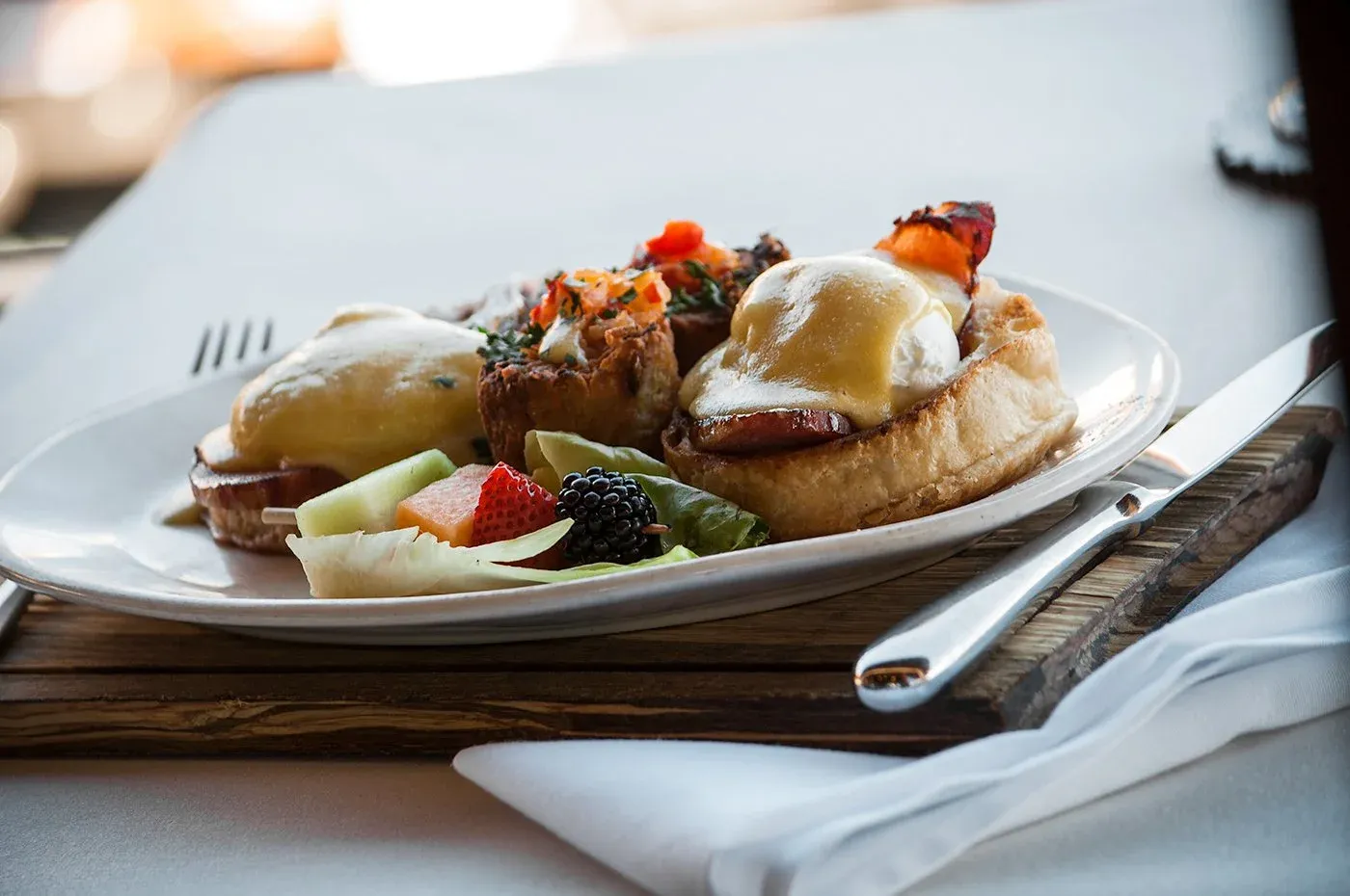 White plate with eggs benedict, savory pastries, and fresh fruit.