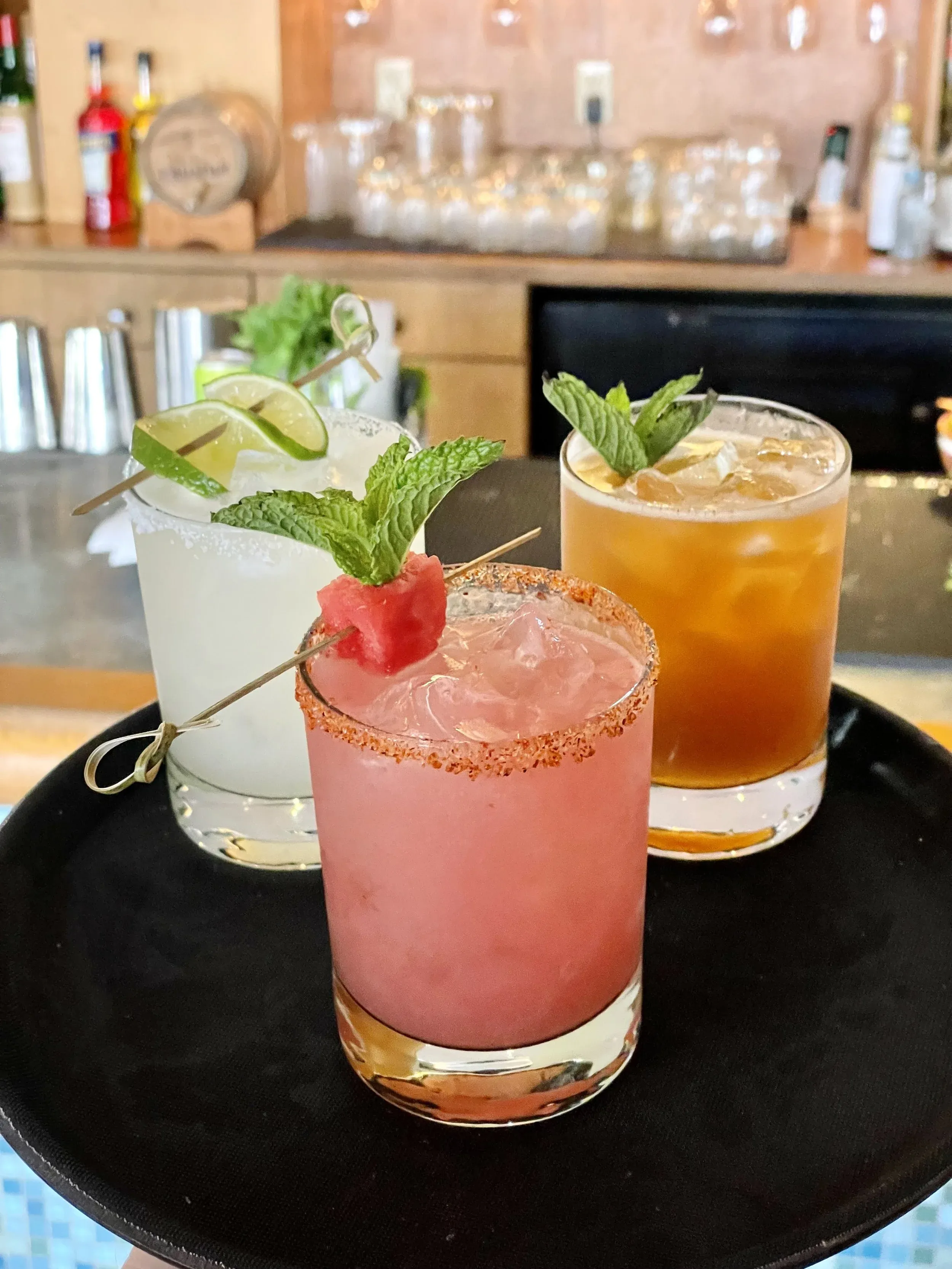Three different cocktails, garnished, served on a black tray.