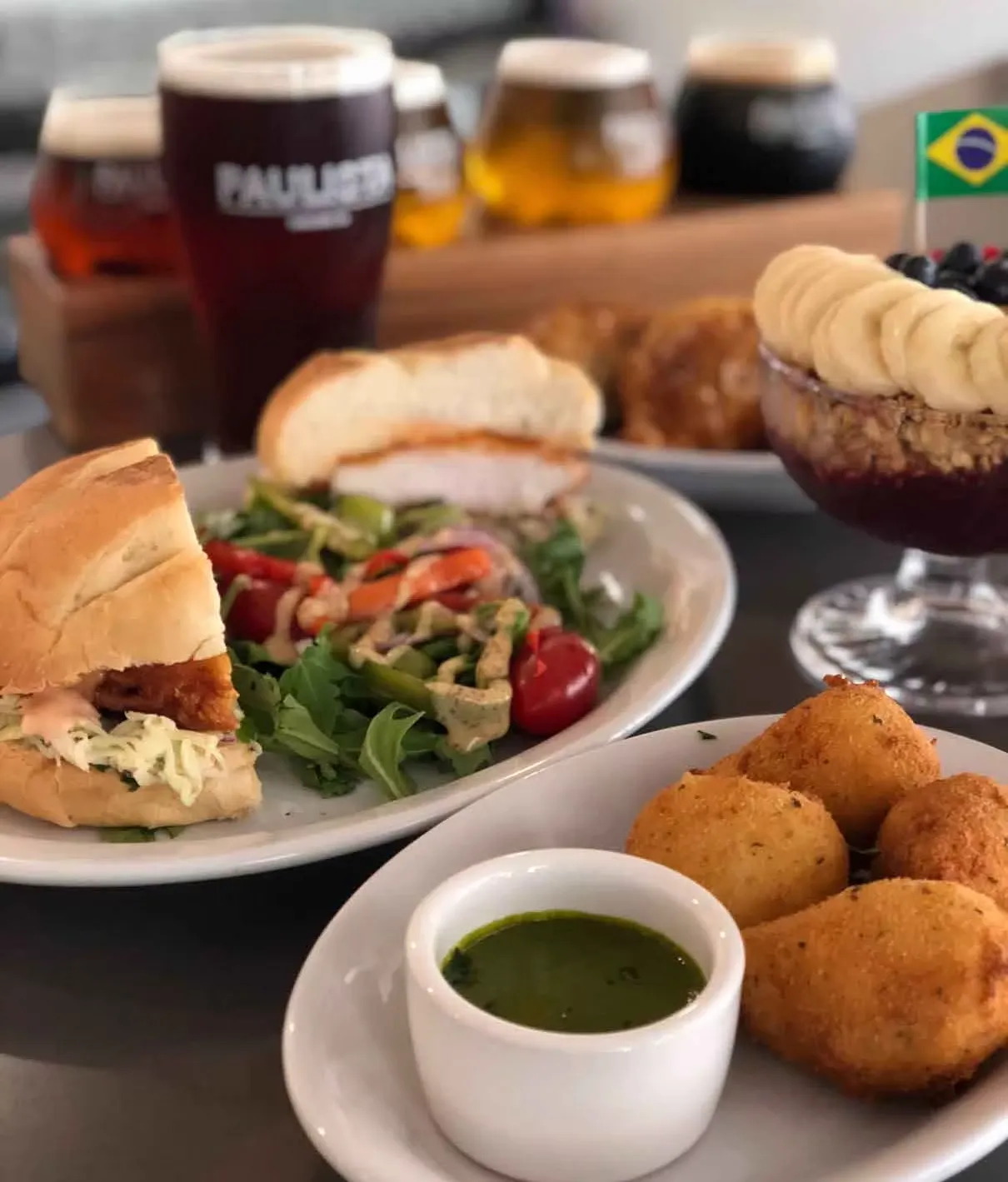 Brazilian meal spread with croquettes, sandwich, salad, acai, and beers.