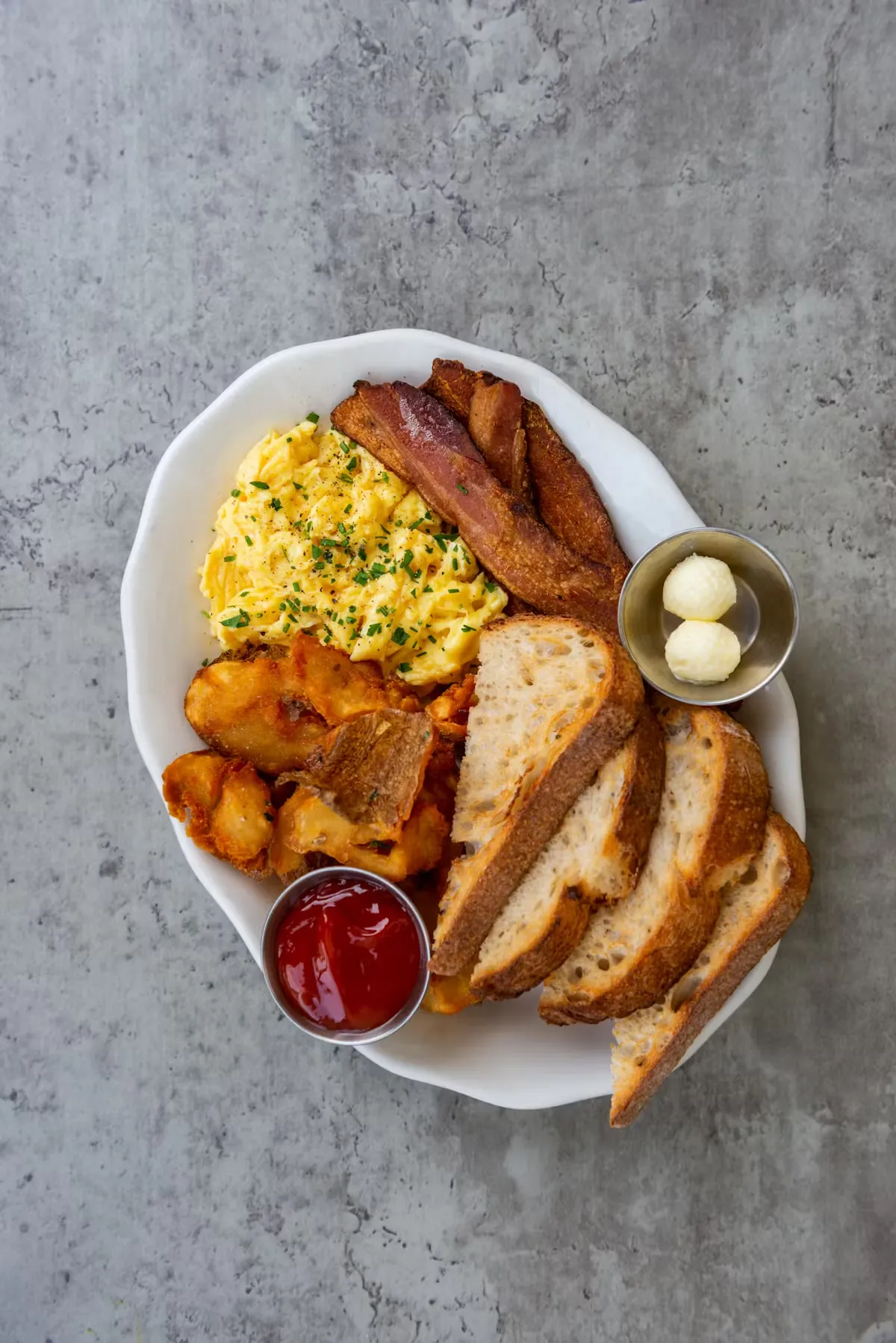 Plate of scrambled eggs, bacon, fried potatoes, toast, butter, and ketchup.