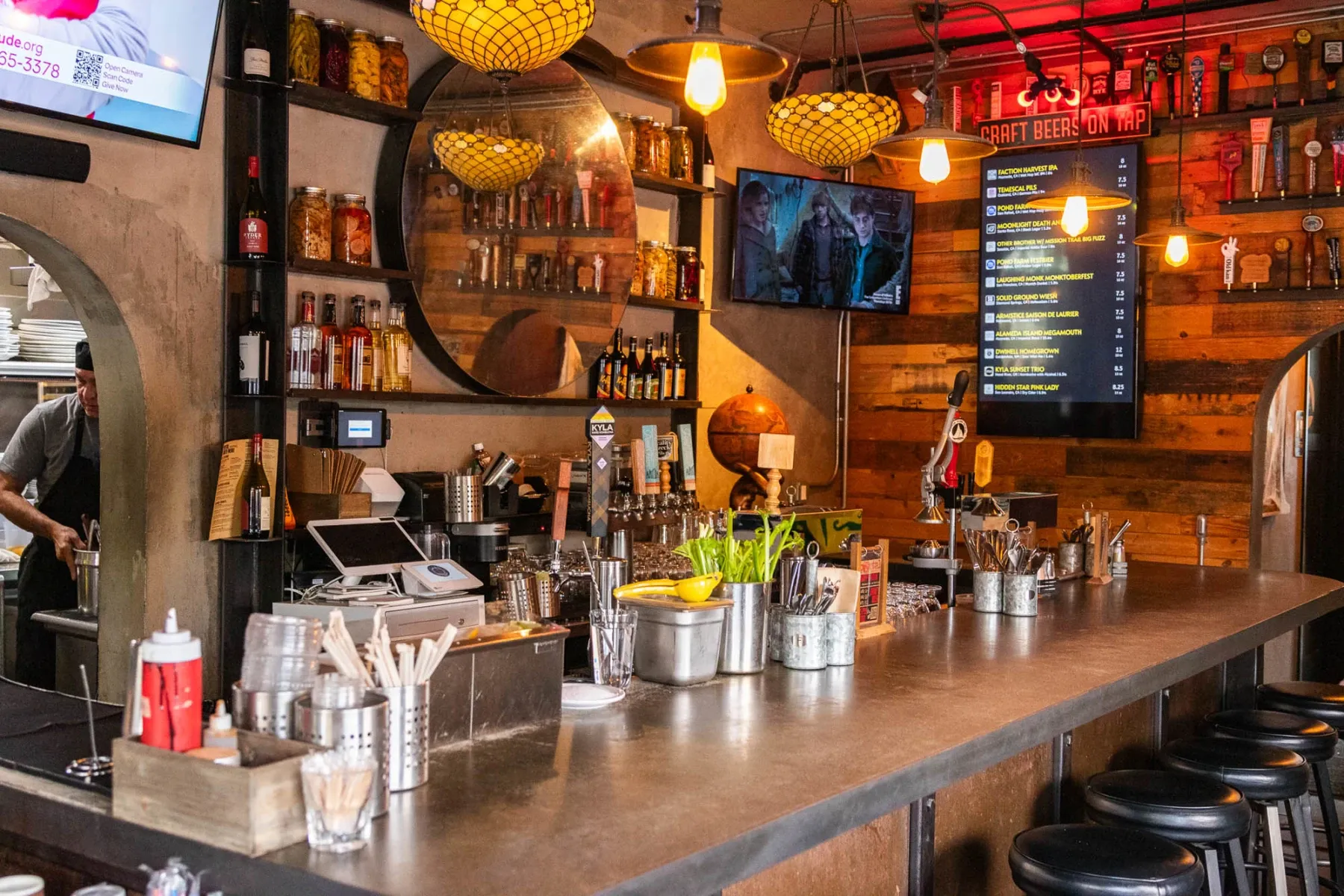 Interior view of a rustic bar with a bartender and menu screens.