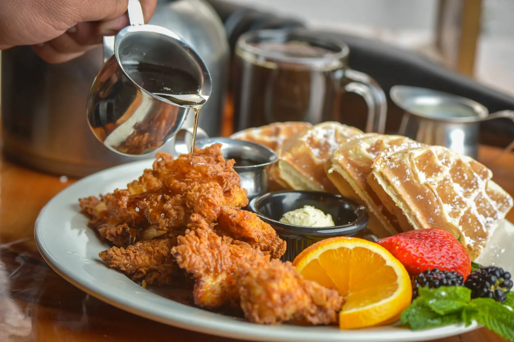 Hand pouring syrup over fried chicken and waffles with fresh fruit.