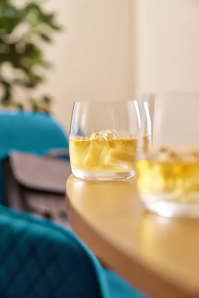 Two tumblers of amber liquid with ice sit on a yellow table.