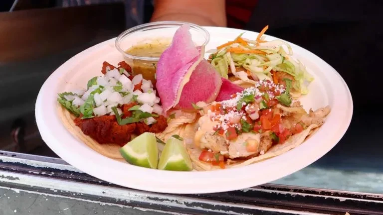 A white plate with two tacos, sauce, watermelon radish, and lime wedges.