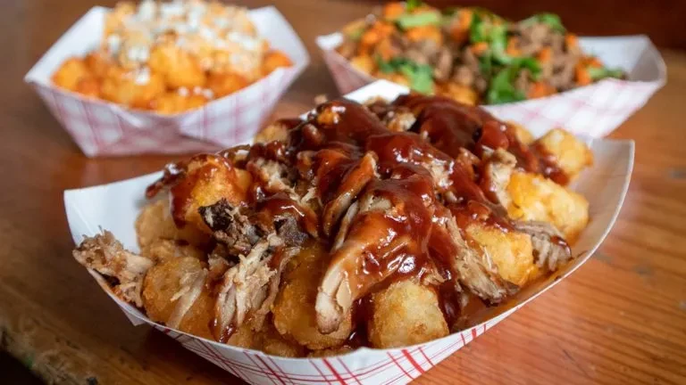 Three paper boats of tater tots with various toppings on a wooden table.
