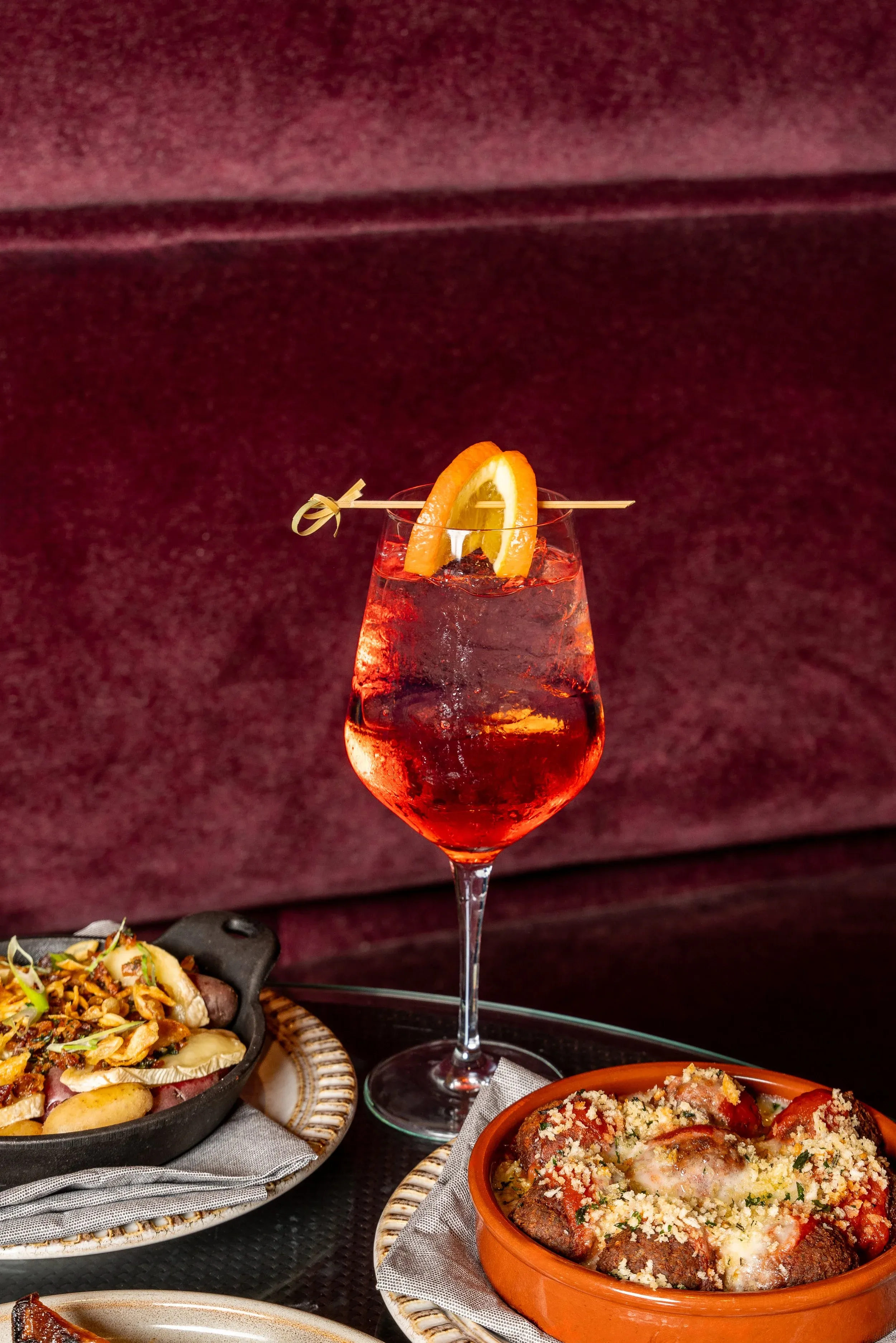 Red cocktail with orange garnish and food on a red velvet background.