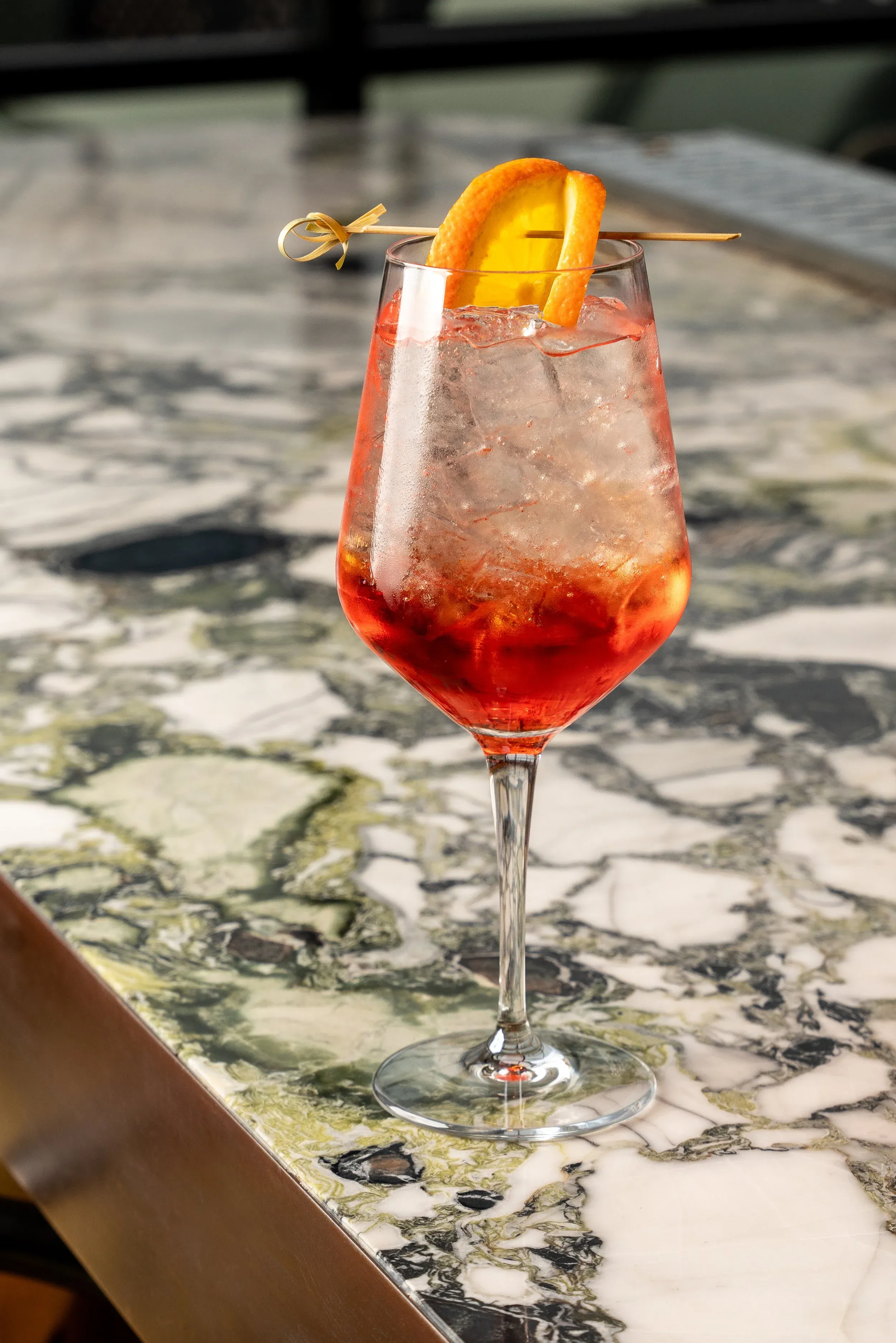 Reddish-orange cocktail with ice and orange slice on marble counter.