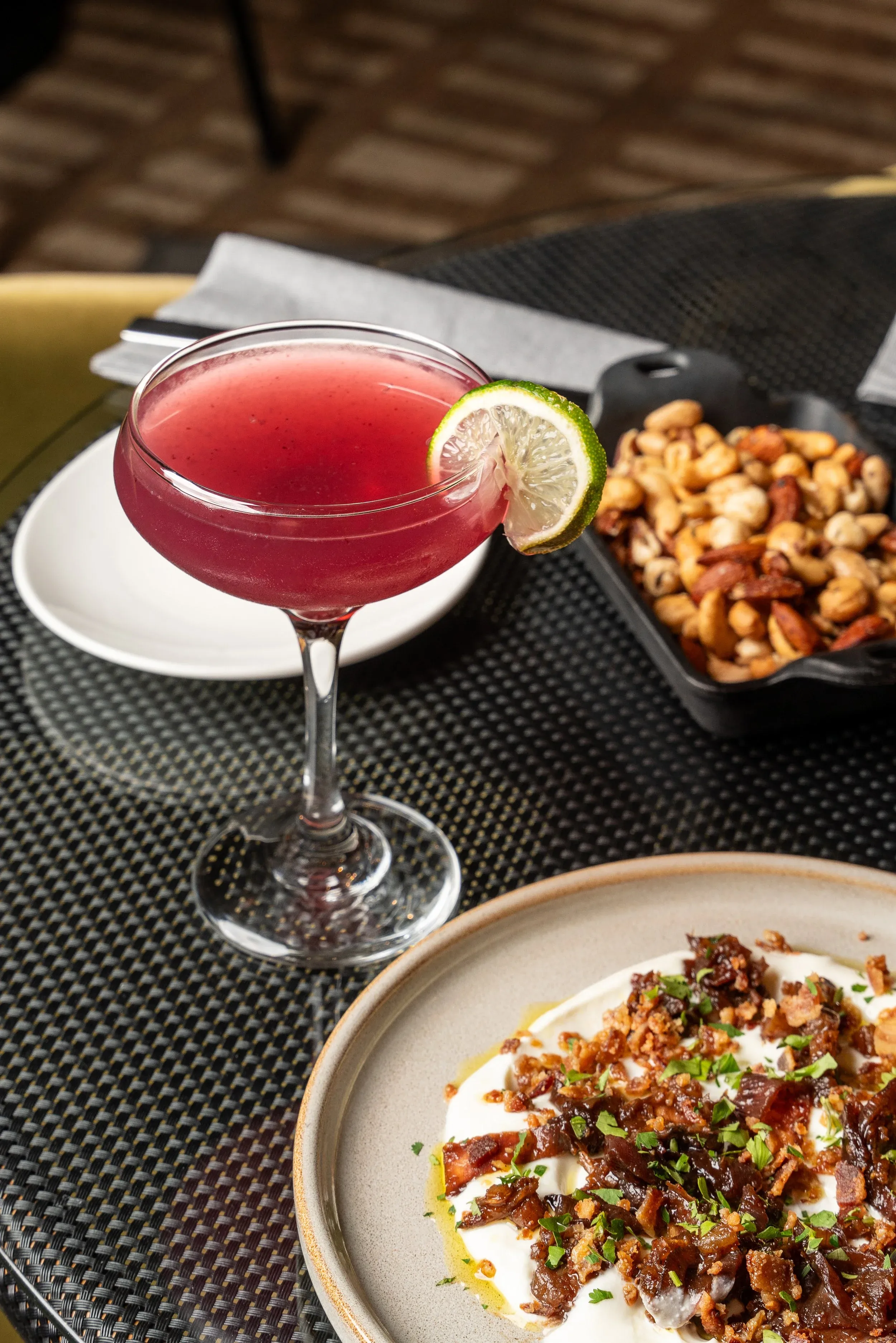 Pink cocktail with lime, mixed nuts, and creamy savory dish.