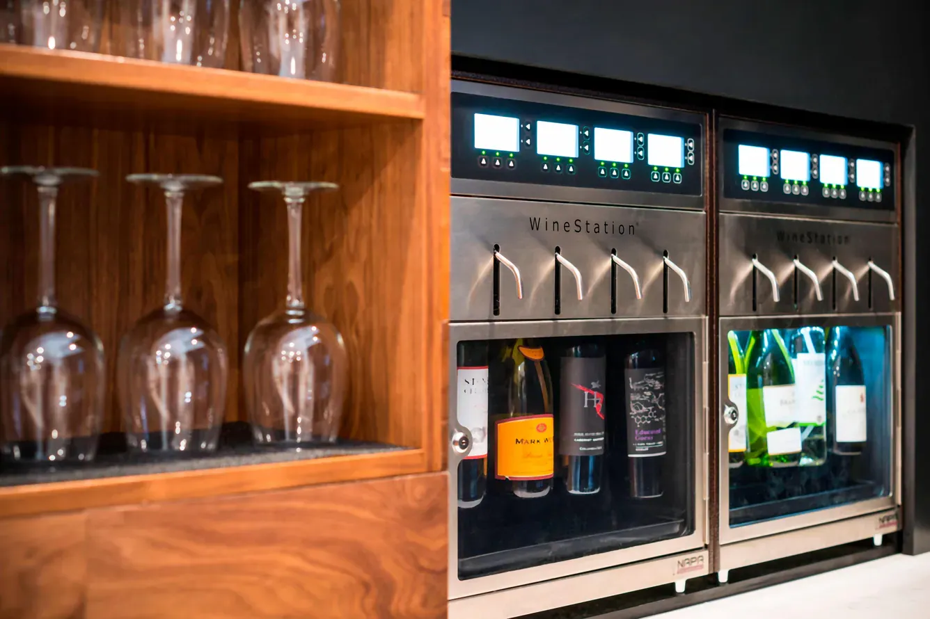 WineStation machines dispensing wine, next to a cabinet with wine glasses.