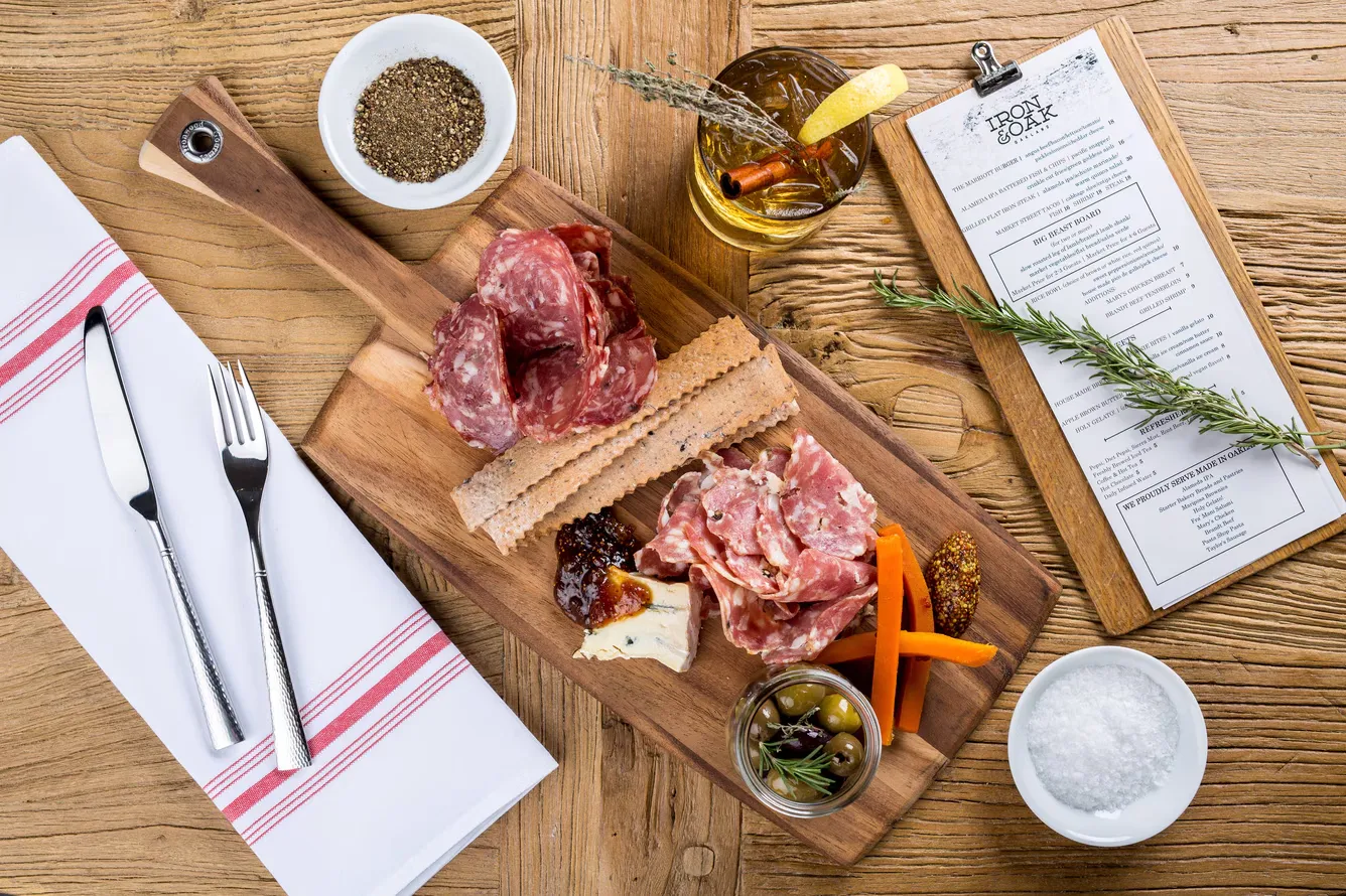 Rustic charcuterie board, cocktail, and menu on a wooden table, overhead.
