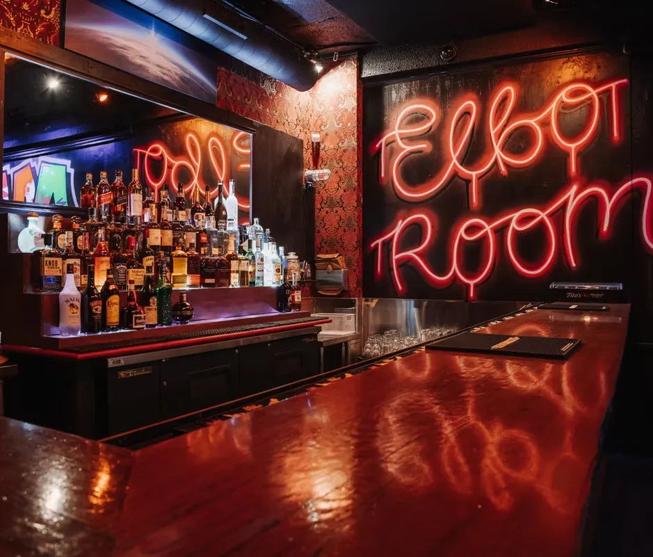 Dark bar interior with red neon "Elbow Room" sign and bottles.