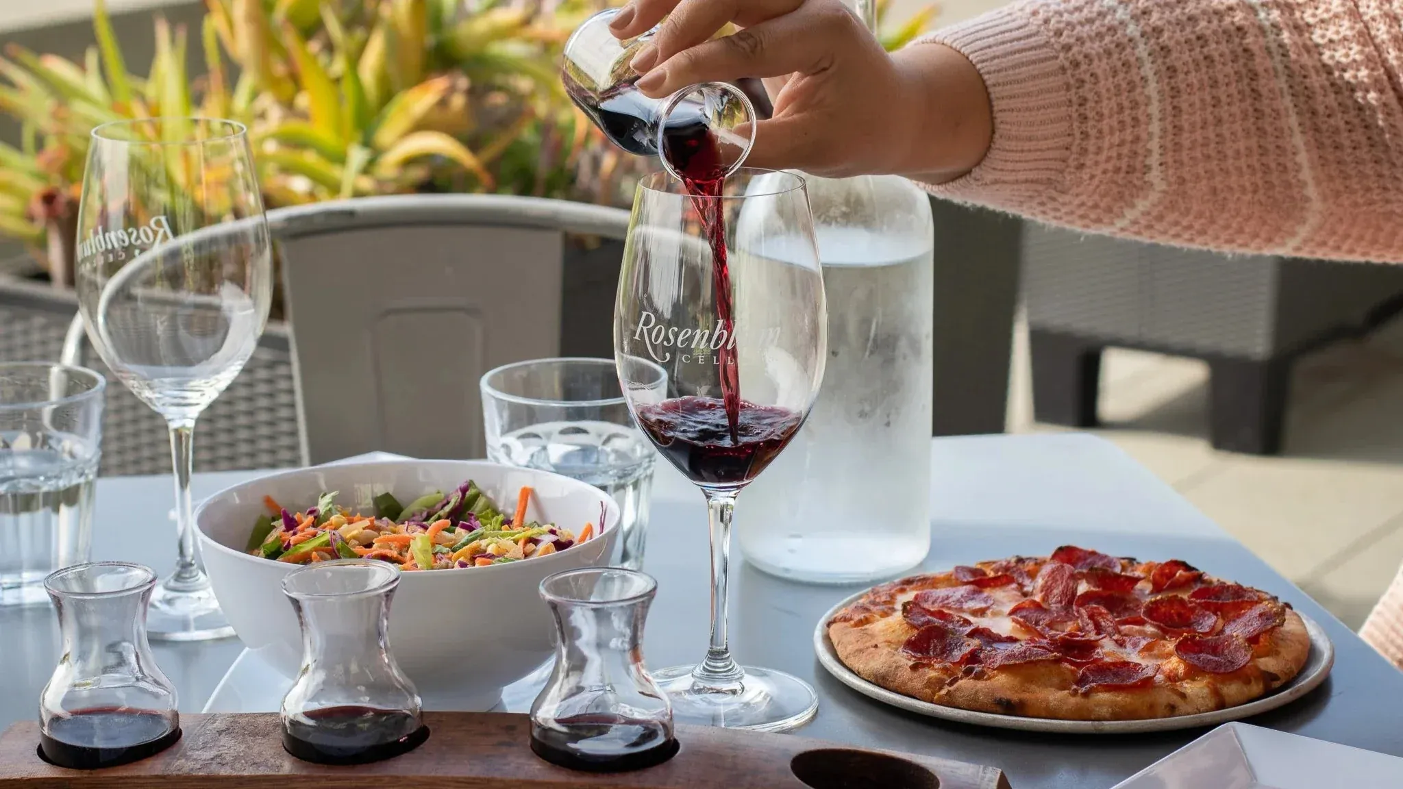 Pouring red wine into a glass at an outdoor table with food.