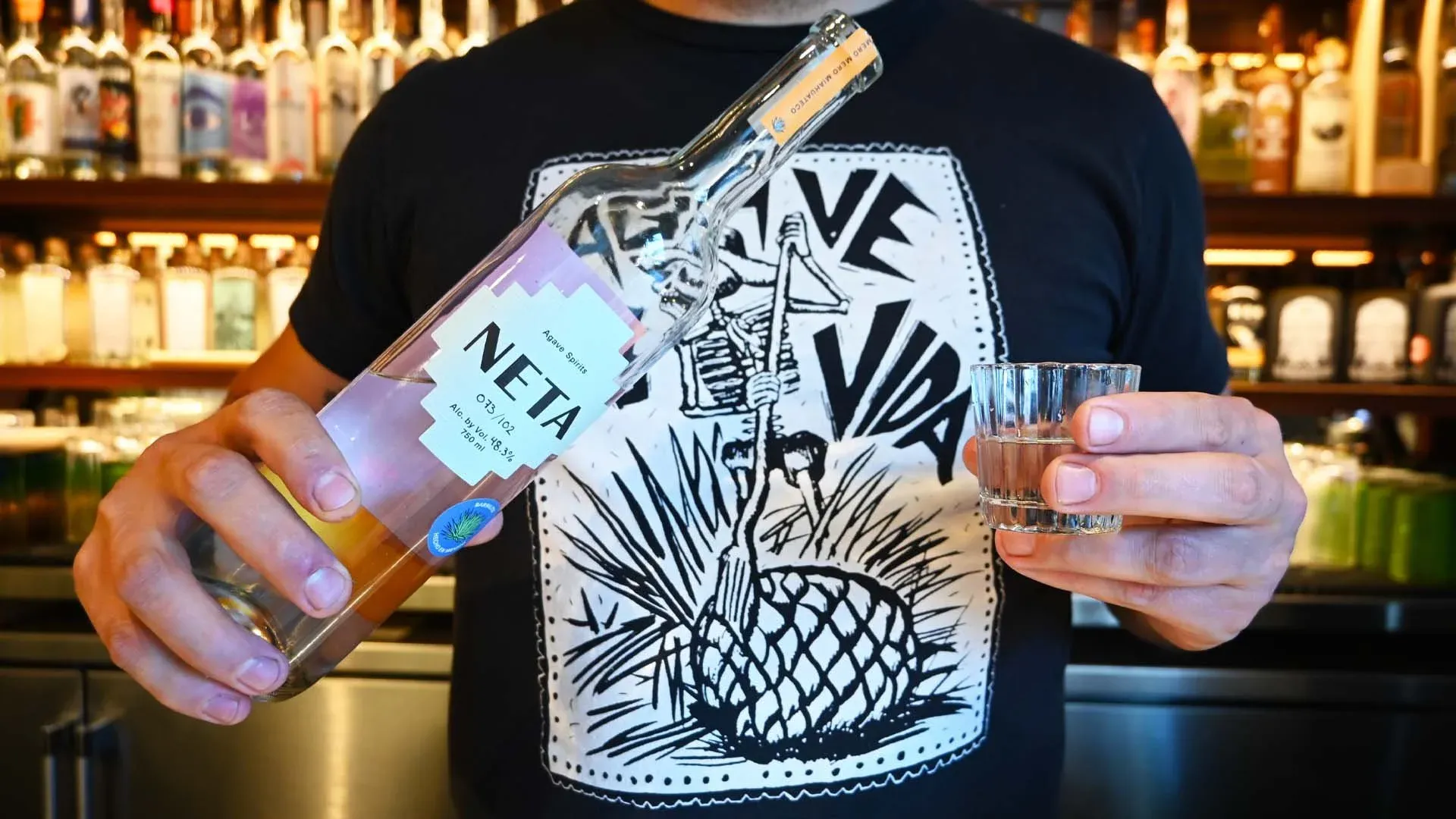 Person holding NETA Agave Spirits bottle and a small glass.