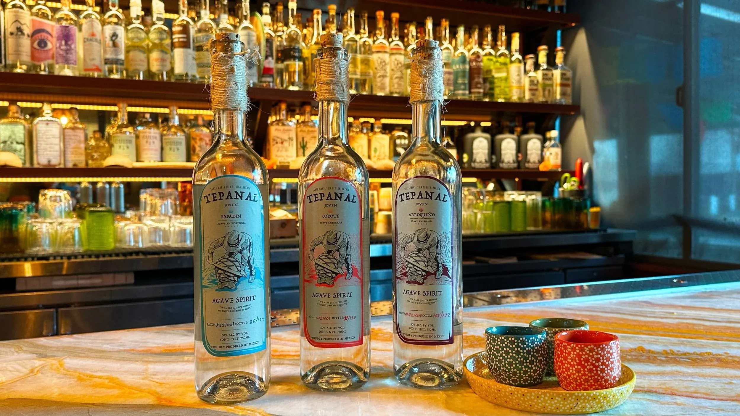 Three agave spirit bottles and colorful shot glasses on a bar.