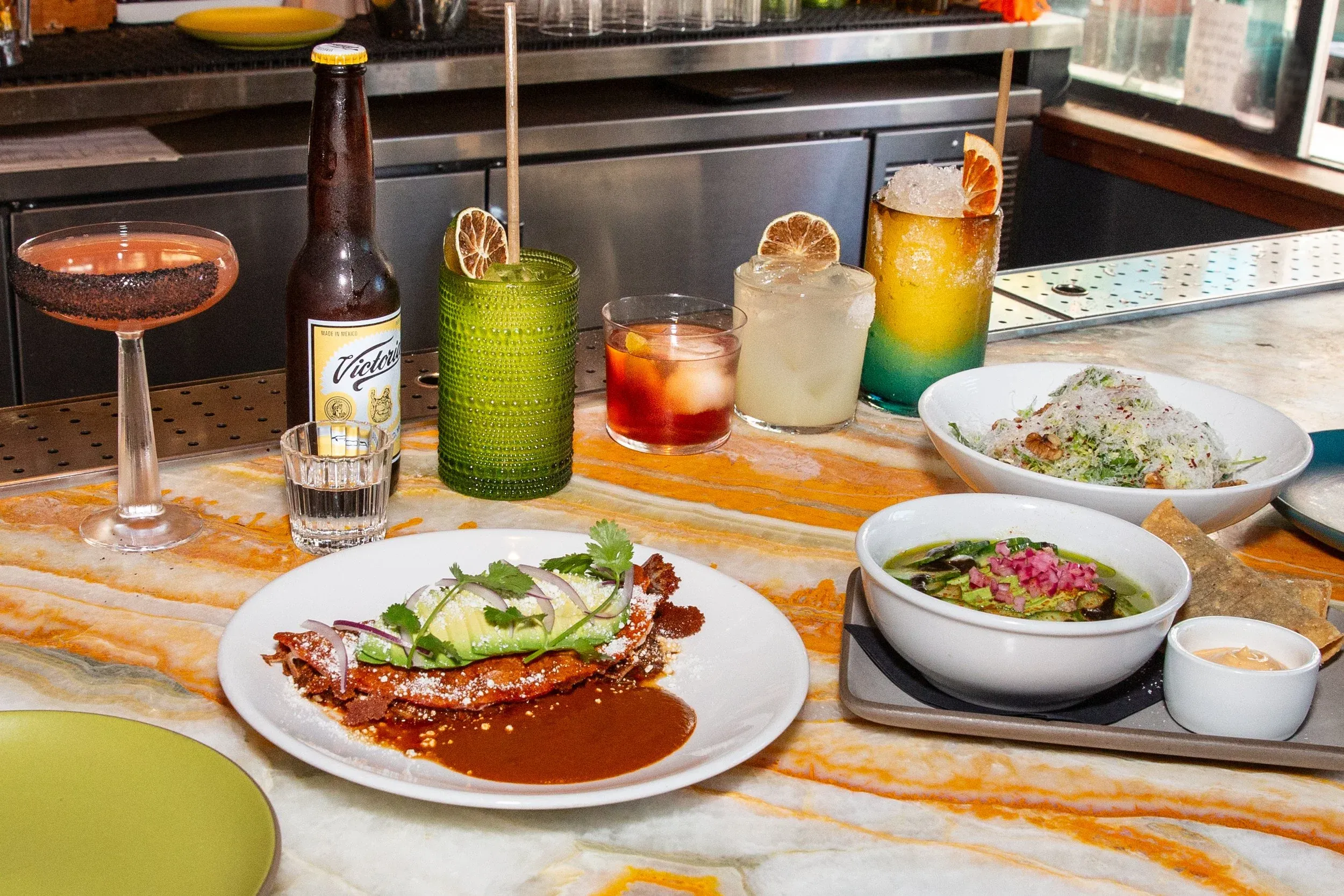 Multiple cocktails, Victoria beer, and Mexican food dishes on a bar counter.