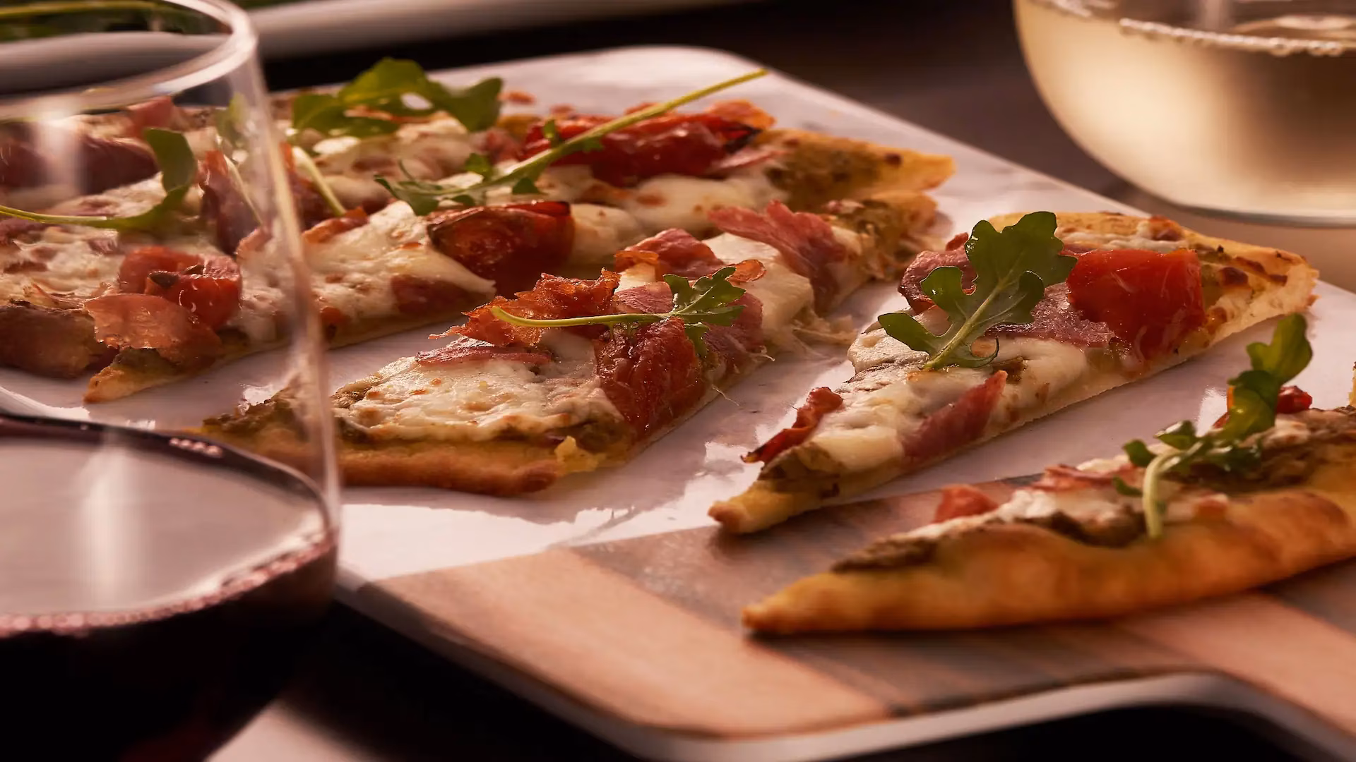 Flatbread slices with cheese, tomatoes, cured meat, arugula, and wine.